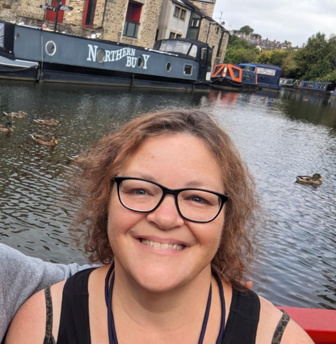 Smiling woman with glasses taking a selfie by a canal with ducks, boats and buildings in the background.
