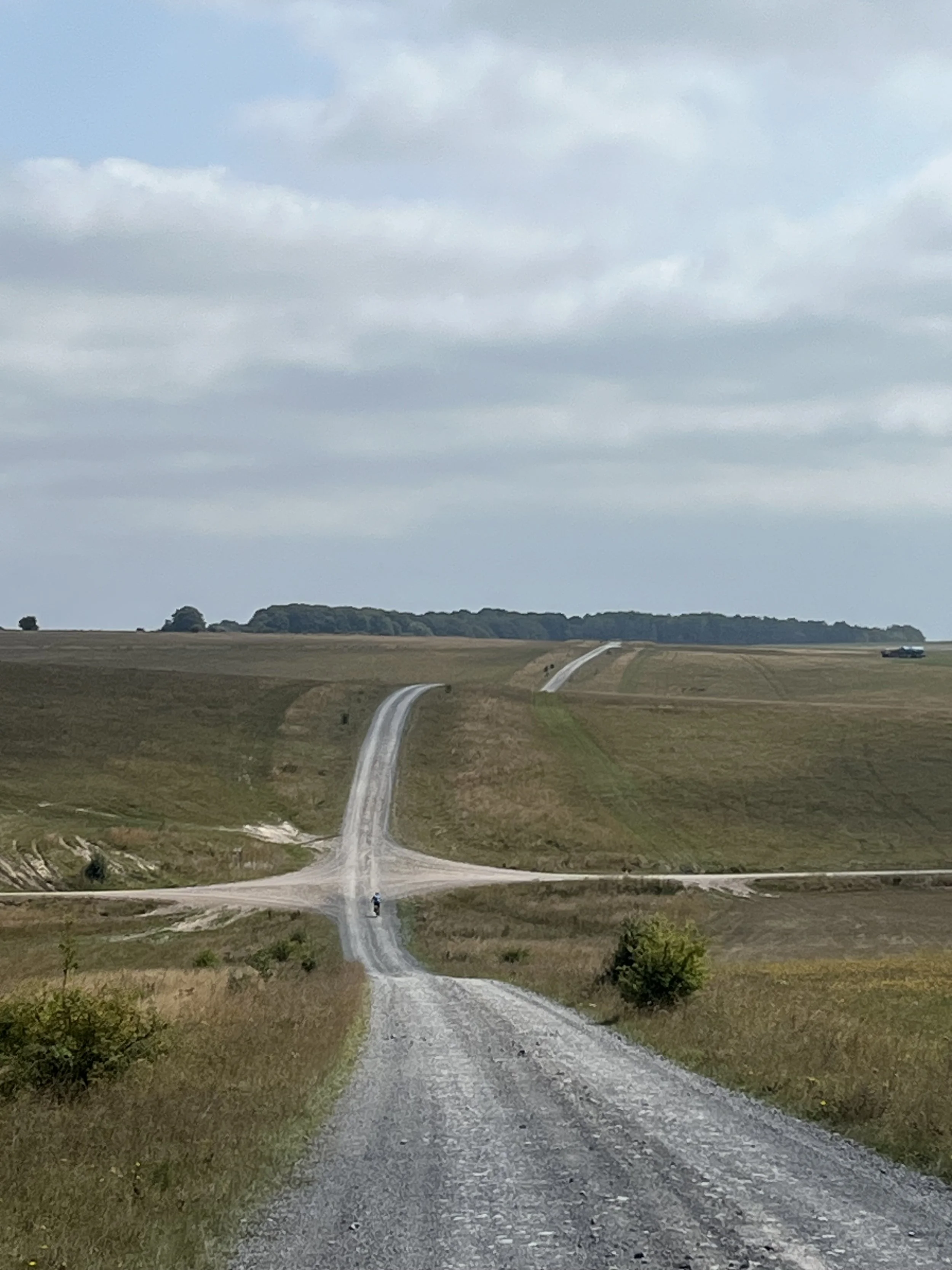 Gravel country road winding through rolling green hills under cloudy sky.