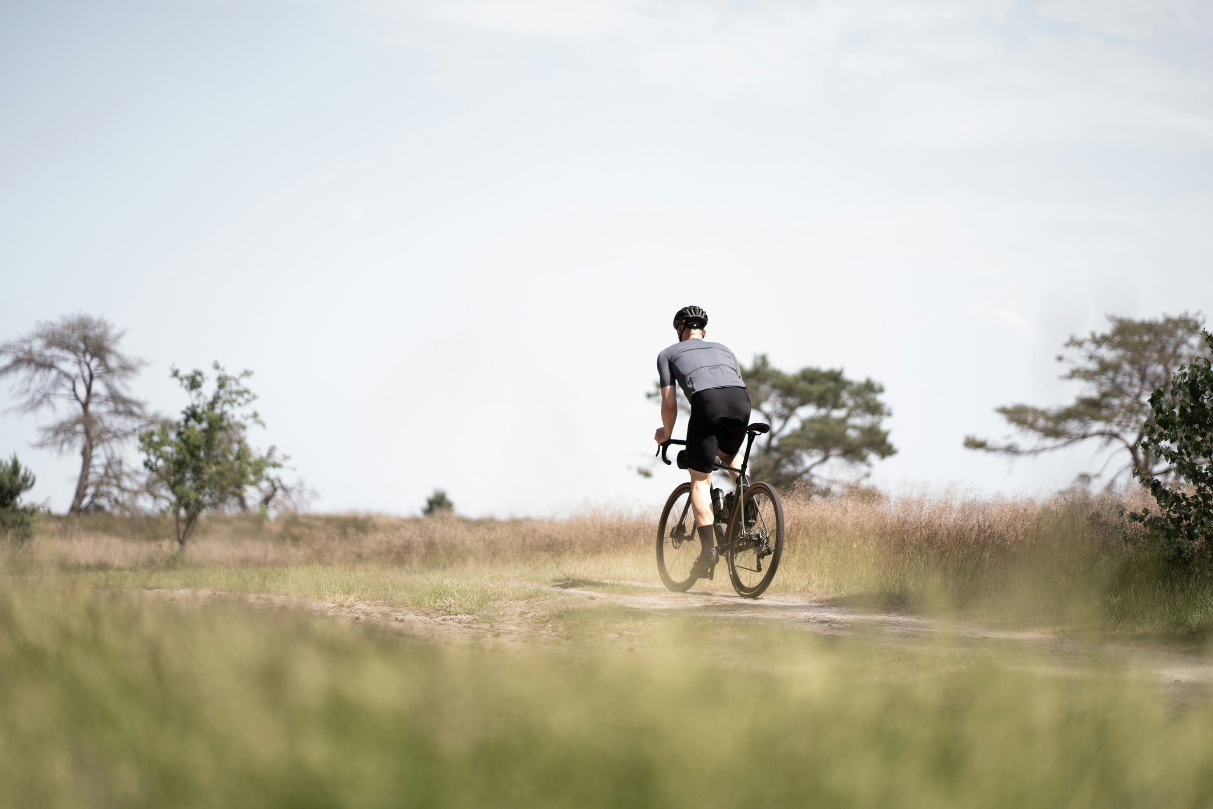 A person riding a bicycle on a trail through a grassy field with trees in the background on a cloudy day.