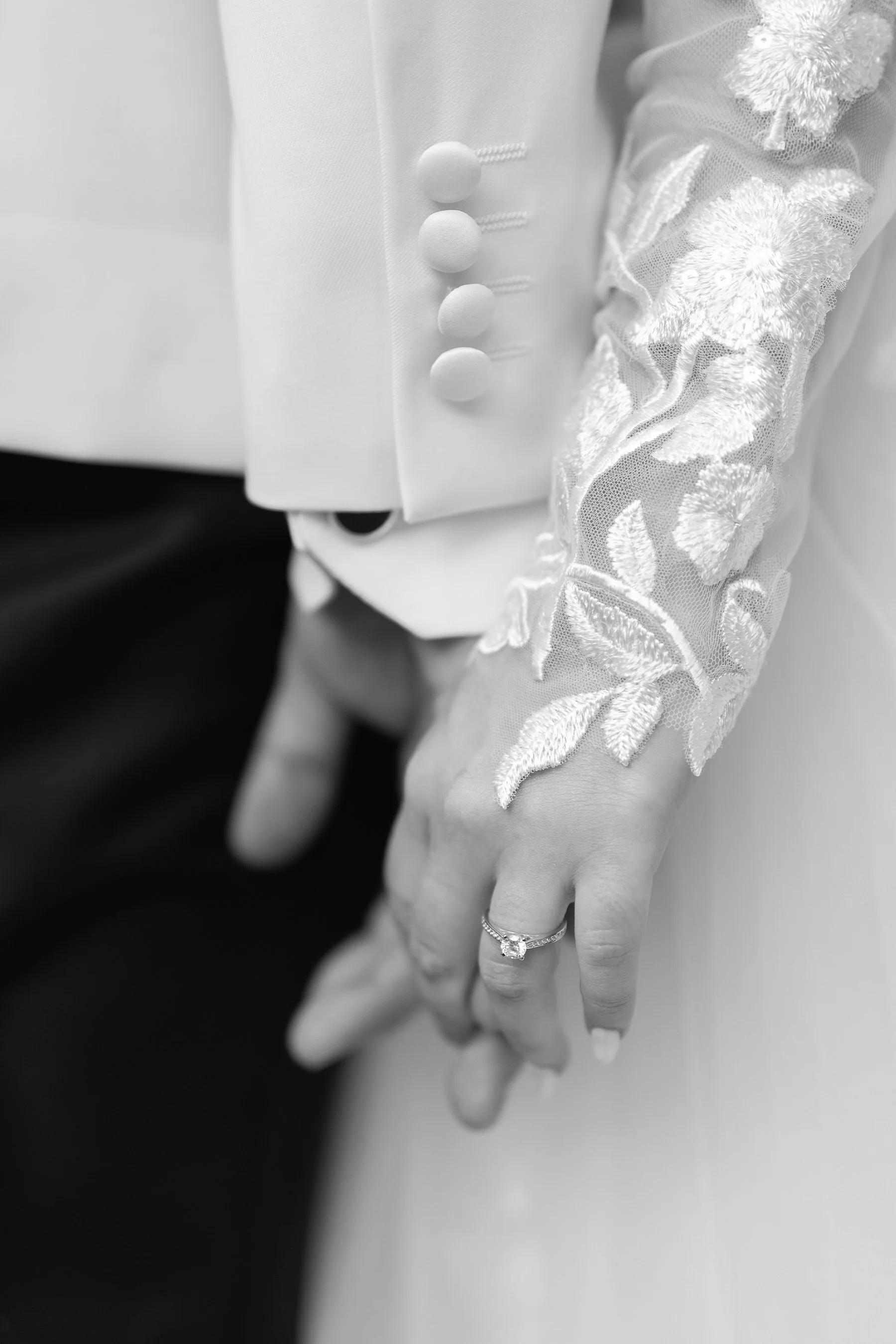 Close-up of a bride and groom holding hands, with the bride’s hand showing an engagement ring, wearing a lace dress and the groom wearing a tuxedo with decorative buttons.