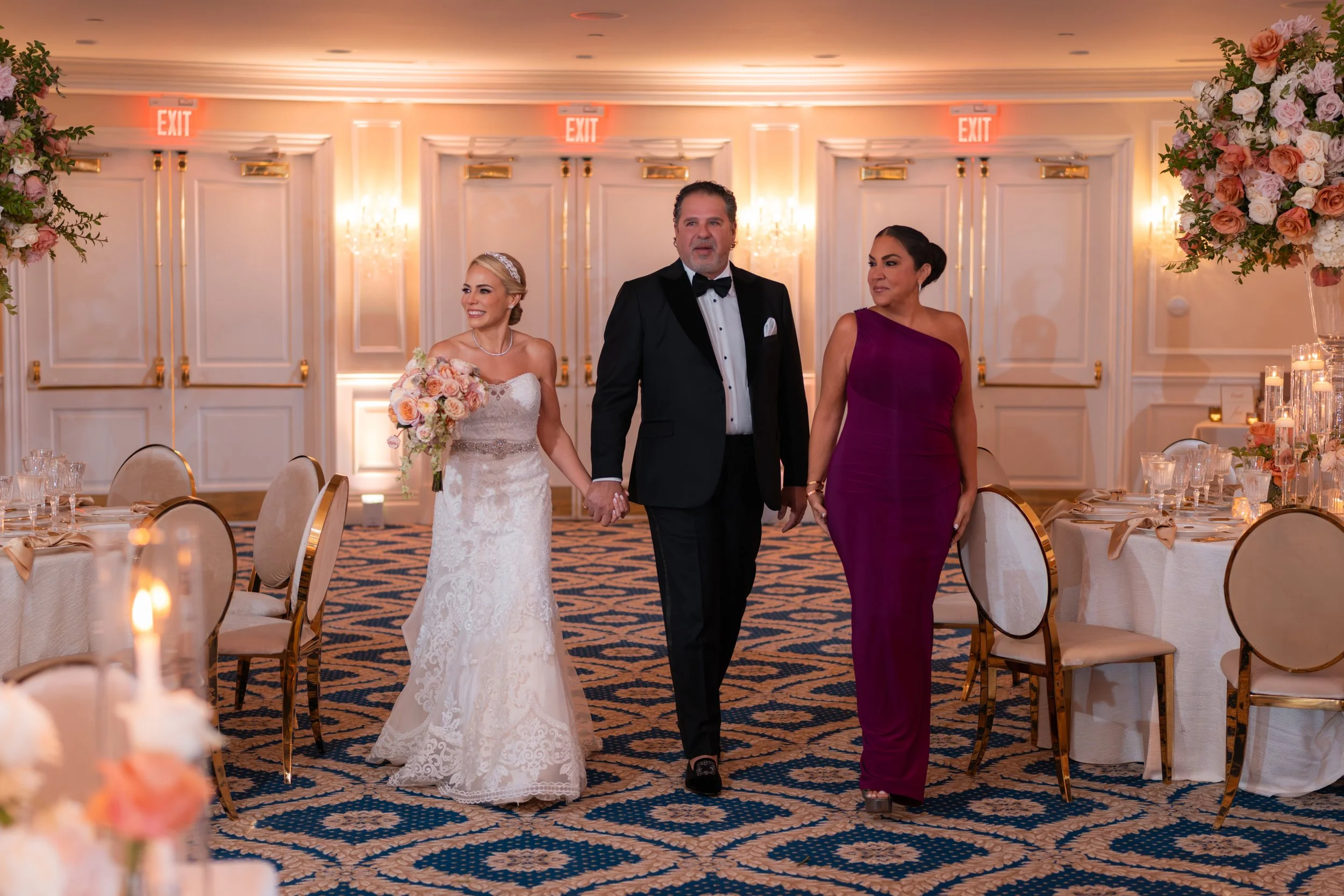A bride, groom, and another woman walking hand-in-hand in a decorated banquet hall. The bride is in a white lace wedding gown holding a bouquet of flowers. The groom is in a black tuxedo with a bow tie. The other woman is dressed in a one-shoulder ma