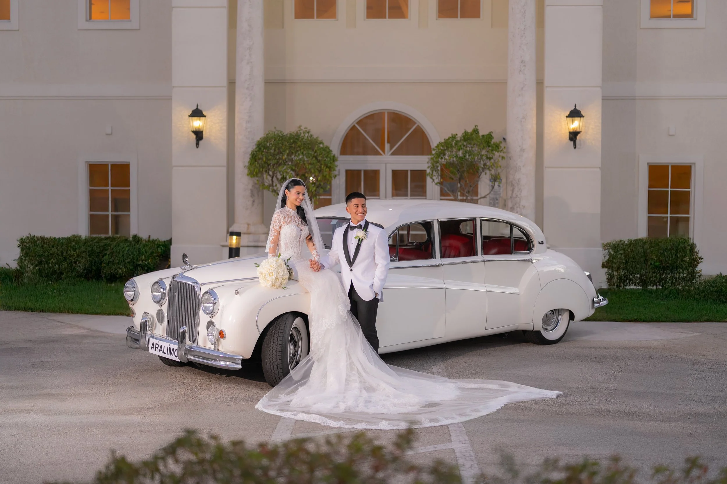 Wedding couple standing beside a vintage white car in front of a building with greenery.