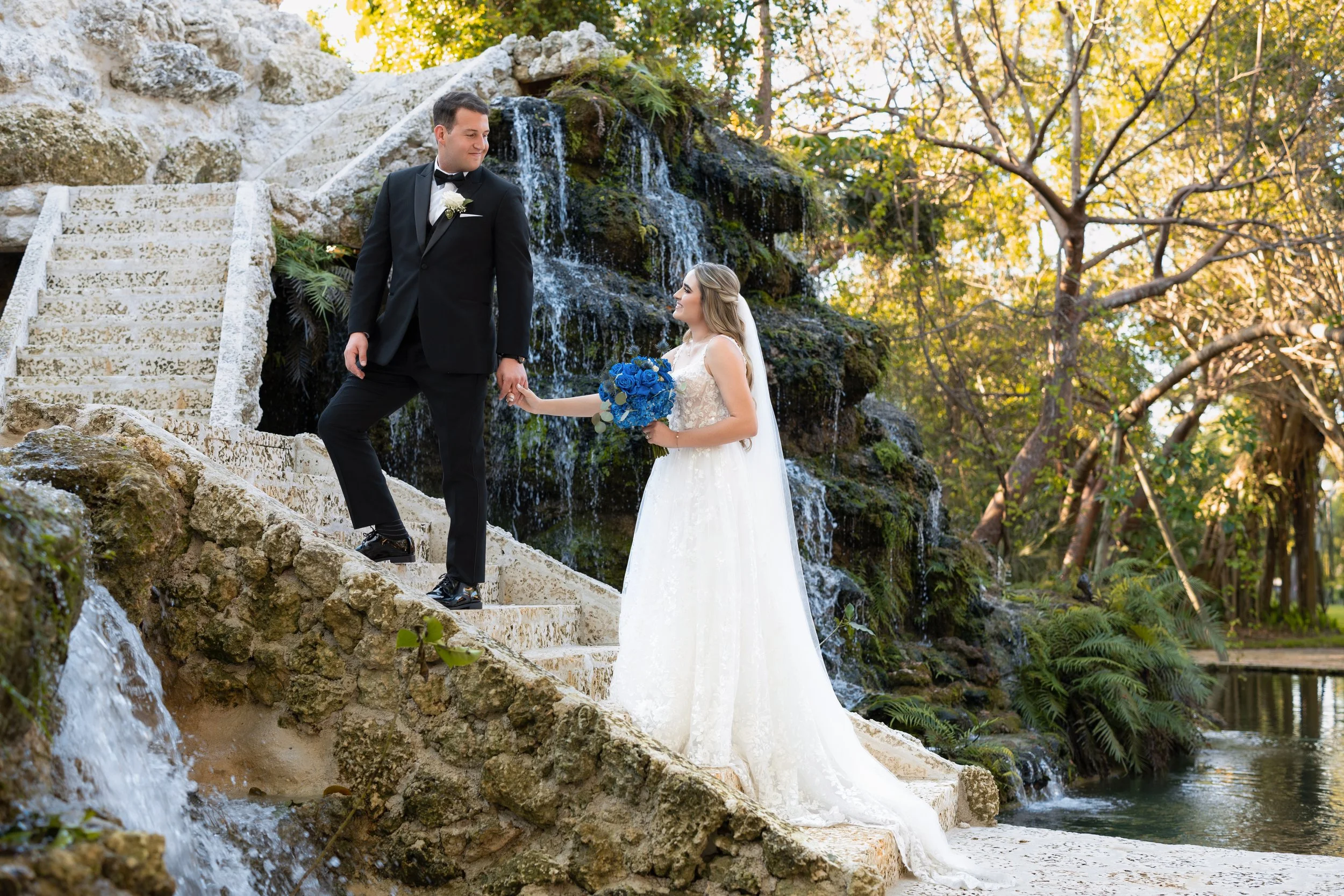 A bride and groom holding hands on a stone staircase beside a waterfall, outdoors in a lush, wooded setting during daytime.