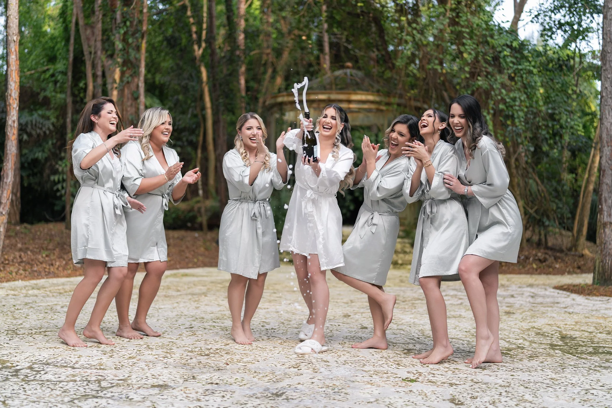 Group of women celebrating outdoors, wearing matching robes, with one opening a bottle of champagne.