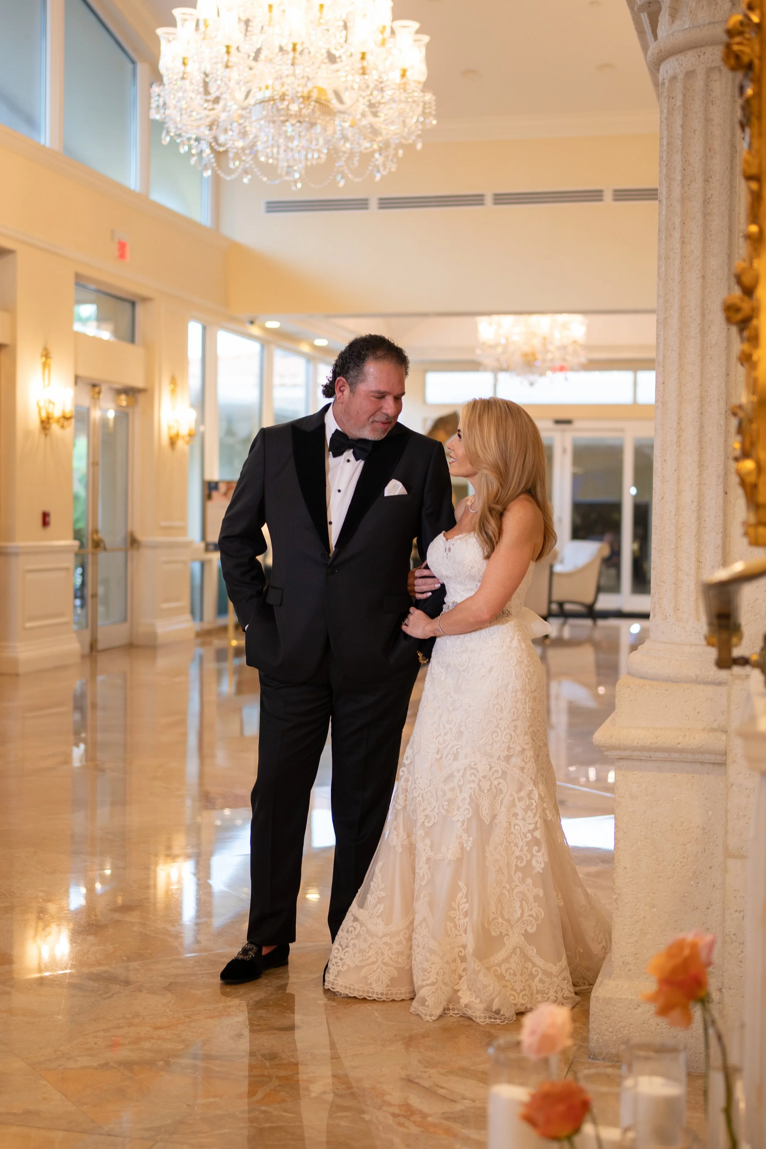 A couple dressed in formal wedding attire, the man in a black tuxedo with a bow tie and the woman in a white lace wedding gown, standing close together and looking at each other in an elegant, well-lit reception hall with chandeliers and floral decor