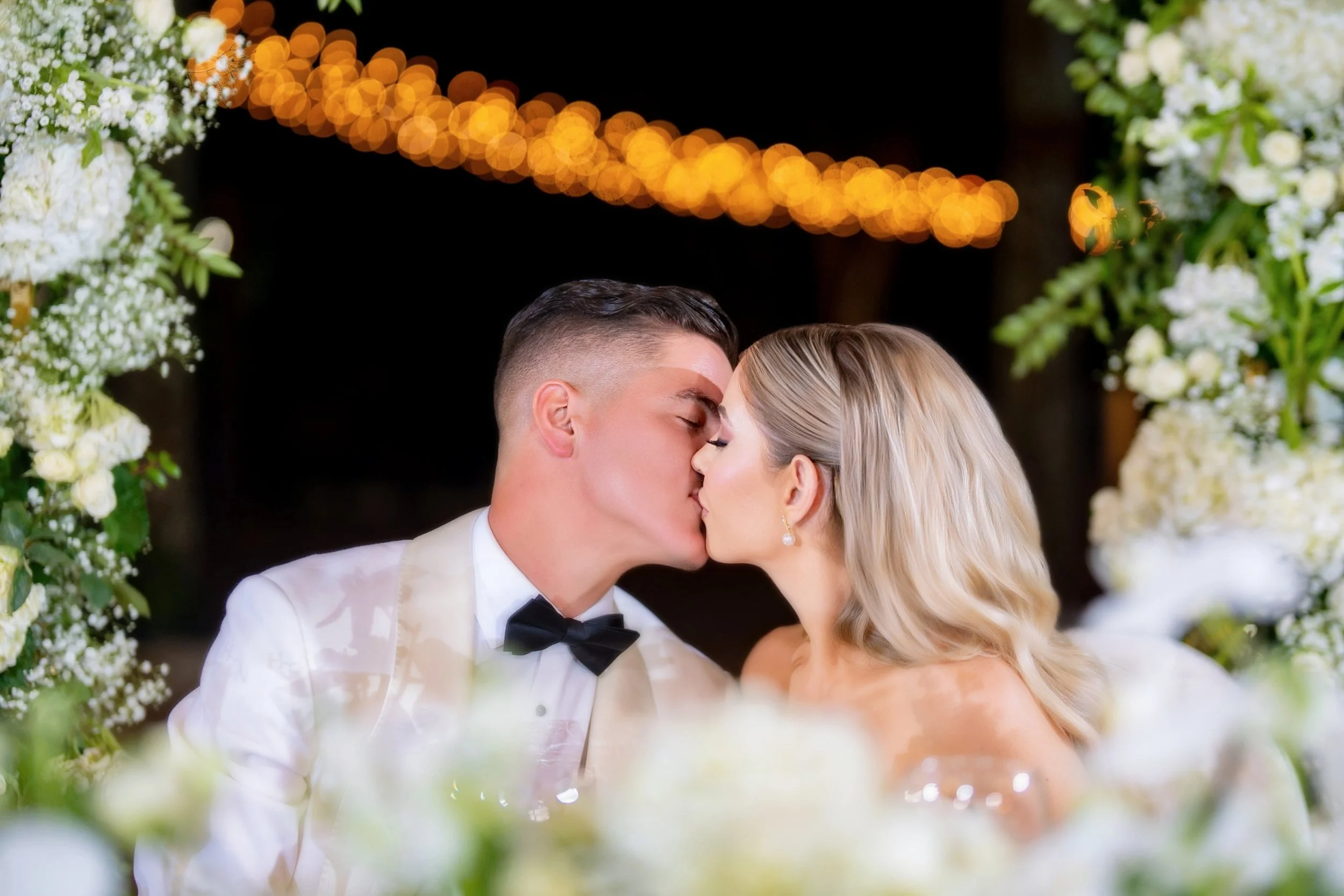 A bride and groom sharing a kiss at their wedding, surrounded by floral arrangements and soft lighting.