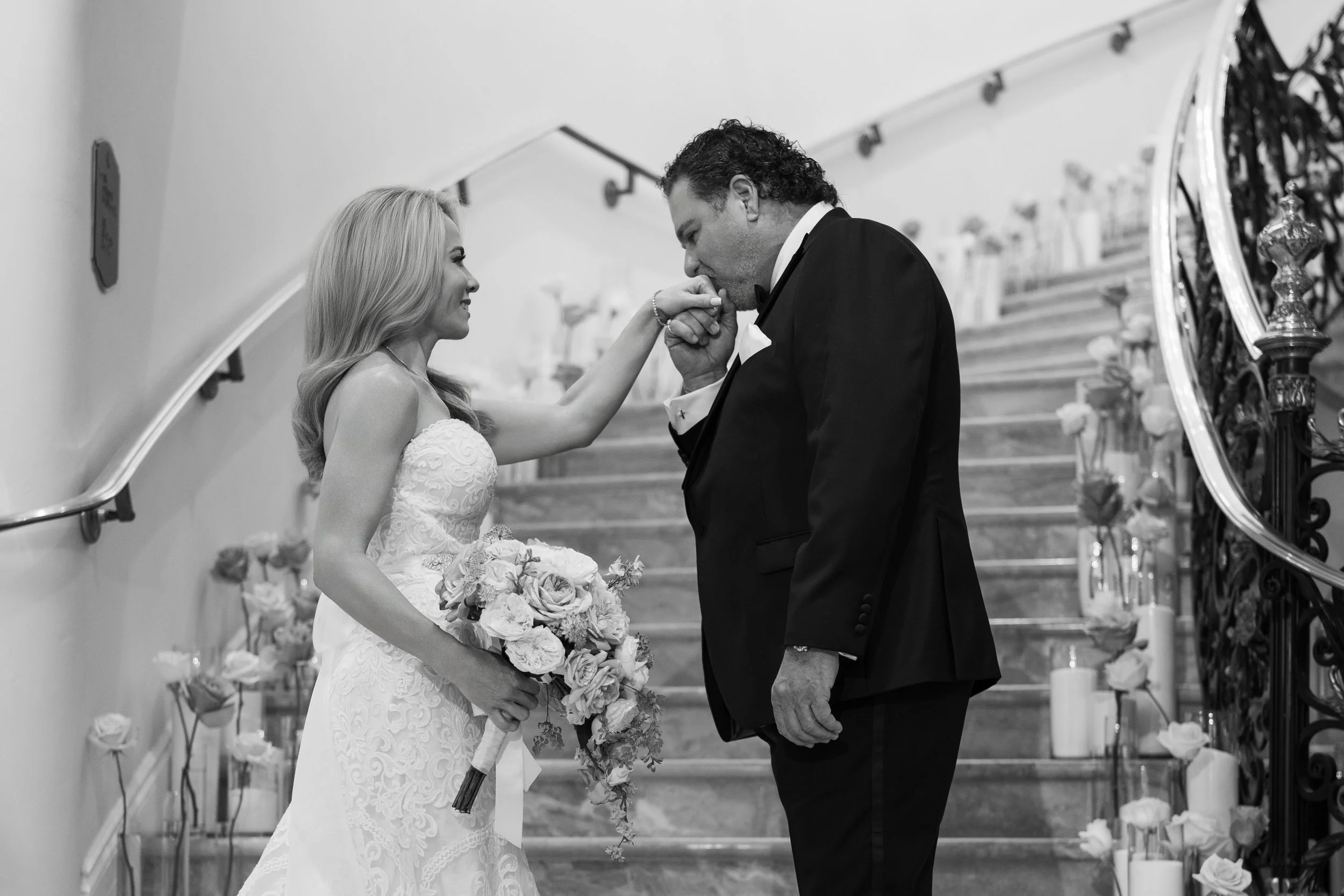 A bride and groom on a staircase, with the groom kissing the bride's hand. The bride holds a bouquet of flowers and wears a lace wedding gown, while the groom wears a black tuxedo.