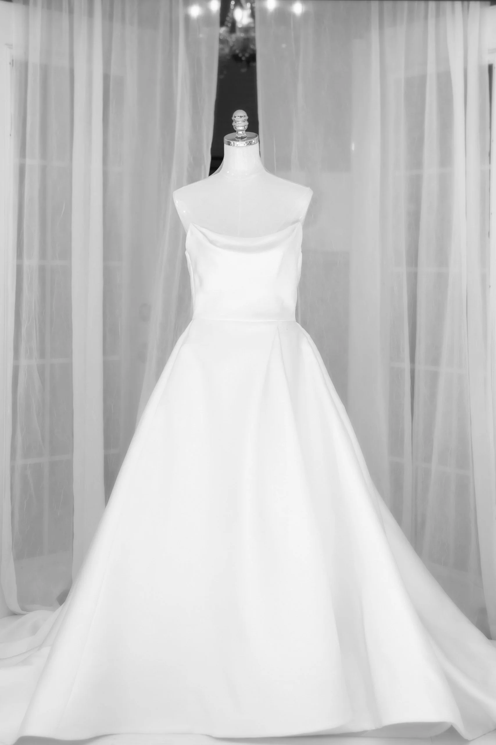A sleeveless white wedding dress displayed on a mannequin in front of sheer curtains.