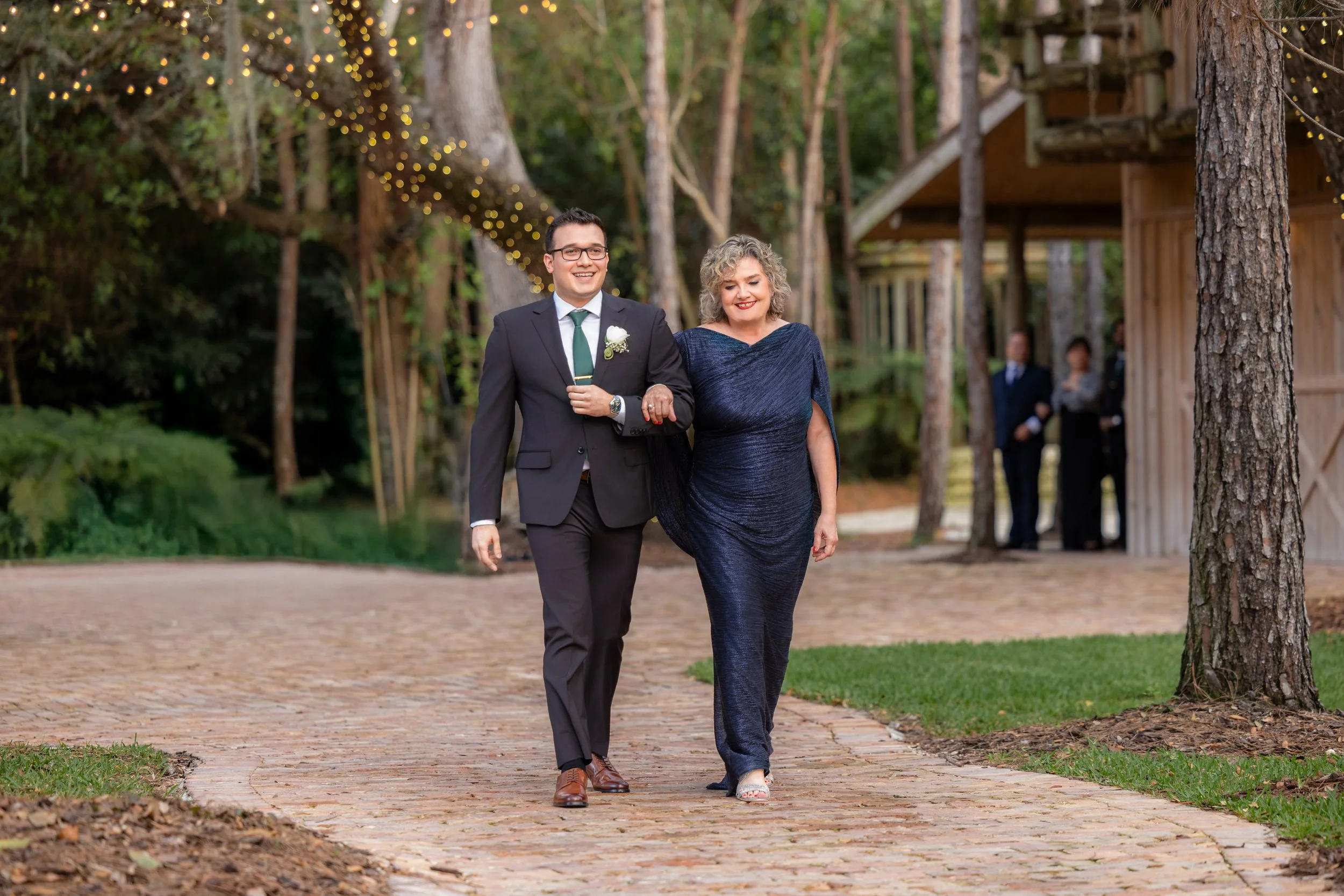 A young man in a black suit and tie walking arm-in-arm with an older woman in a dark blue dress down a brick pathway at an outdoor event, with trees and string lights in the background.