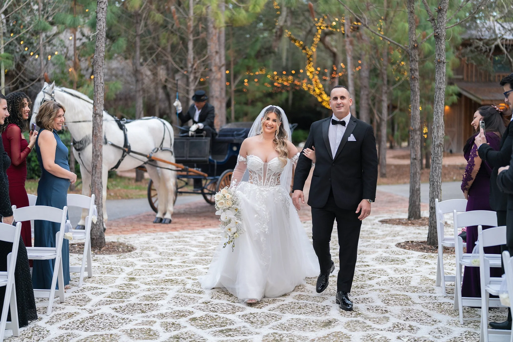A newlywed couple walking down the aisle at an outdoor wedding ceremony, with guests on both sides taking pictures. The bride is in a white wedding gown holding a bouquet, and the groom is in a black tuxedo. There is a horse-drawn carriage in the bac
