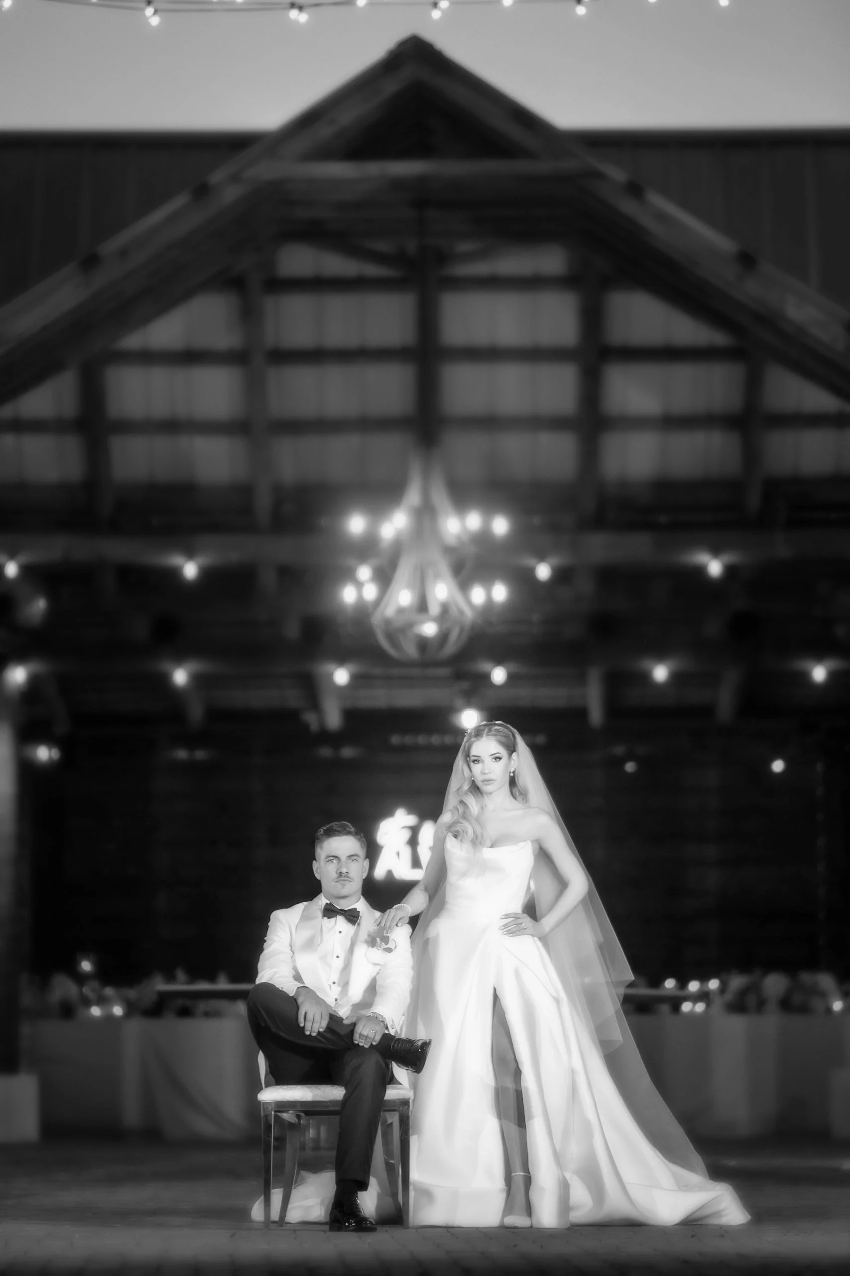 Black and white photo of a bride and groom in wedding attire inside a rustic barn, with the groom sitting on a chair and the bride standing beside him.