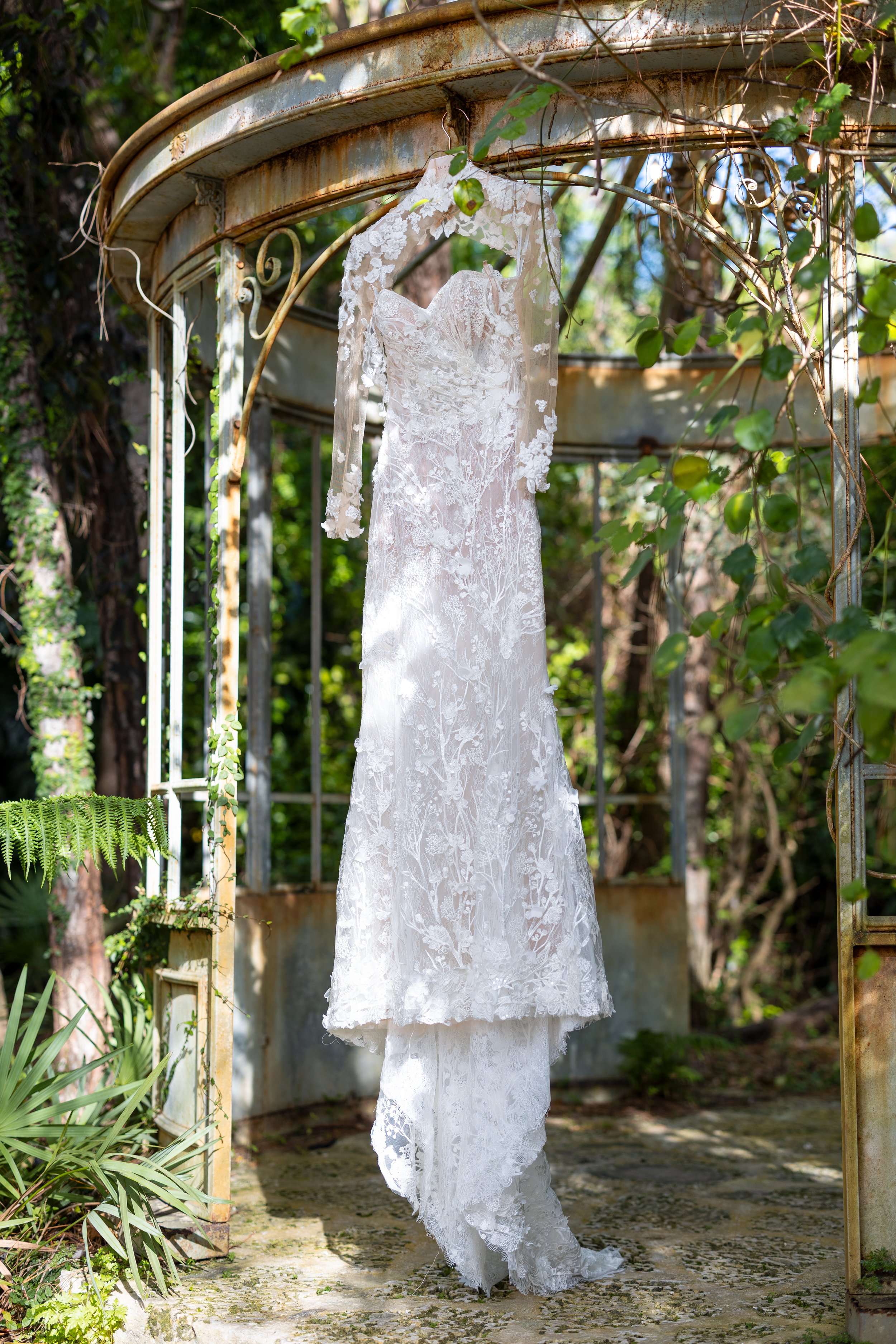 A white lace wedding dress hanging on a metal archway outdoors surrounded by greenery.