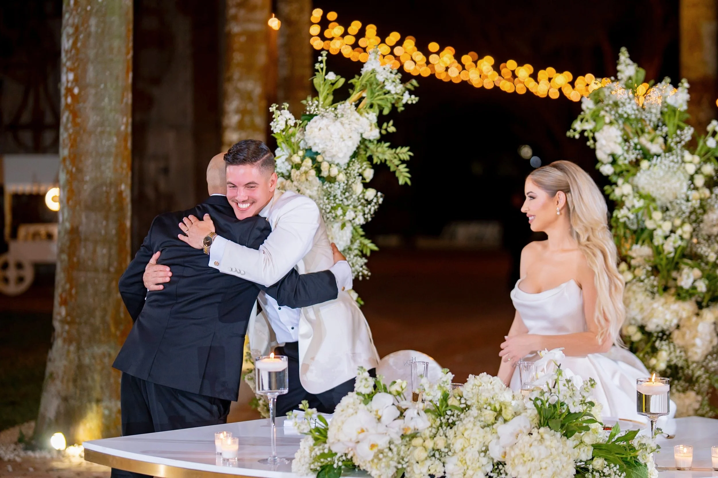 A group of people at a wedding celebration, with two men hugging and a woman smiling, surrounded by white floral decorations and candles.