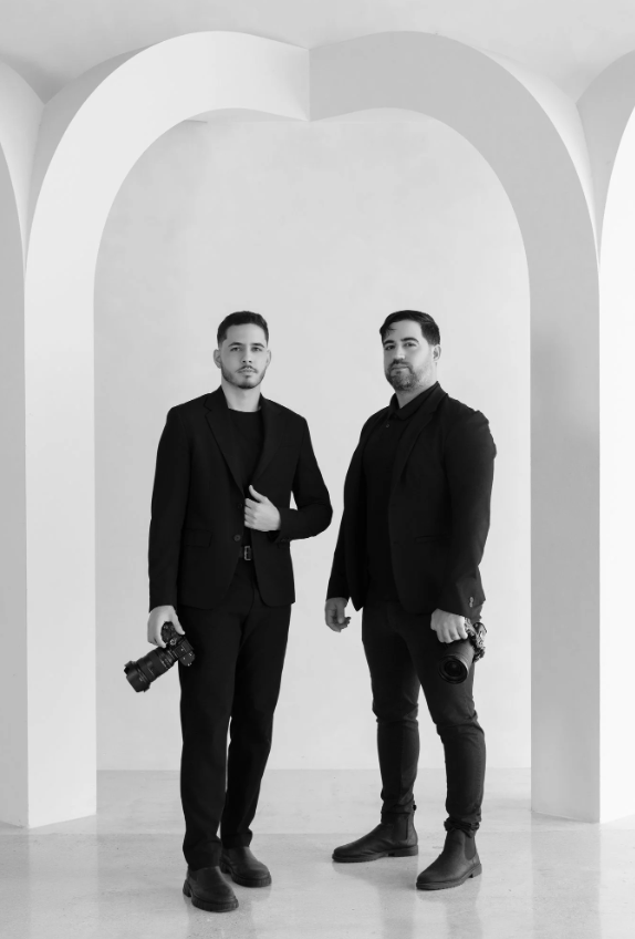 Two men in black suits standing in front of a white archway, each holding a camera.