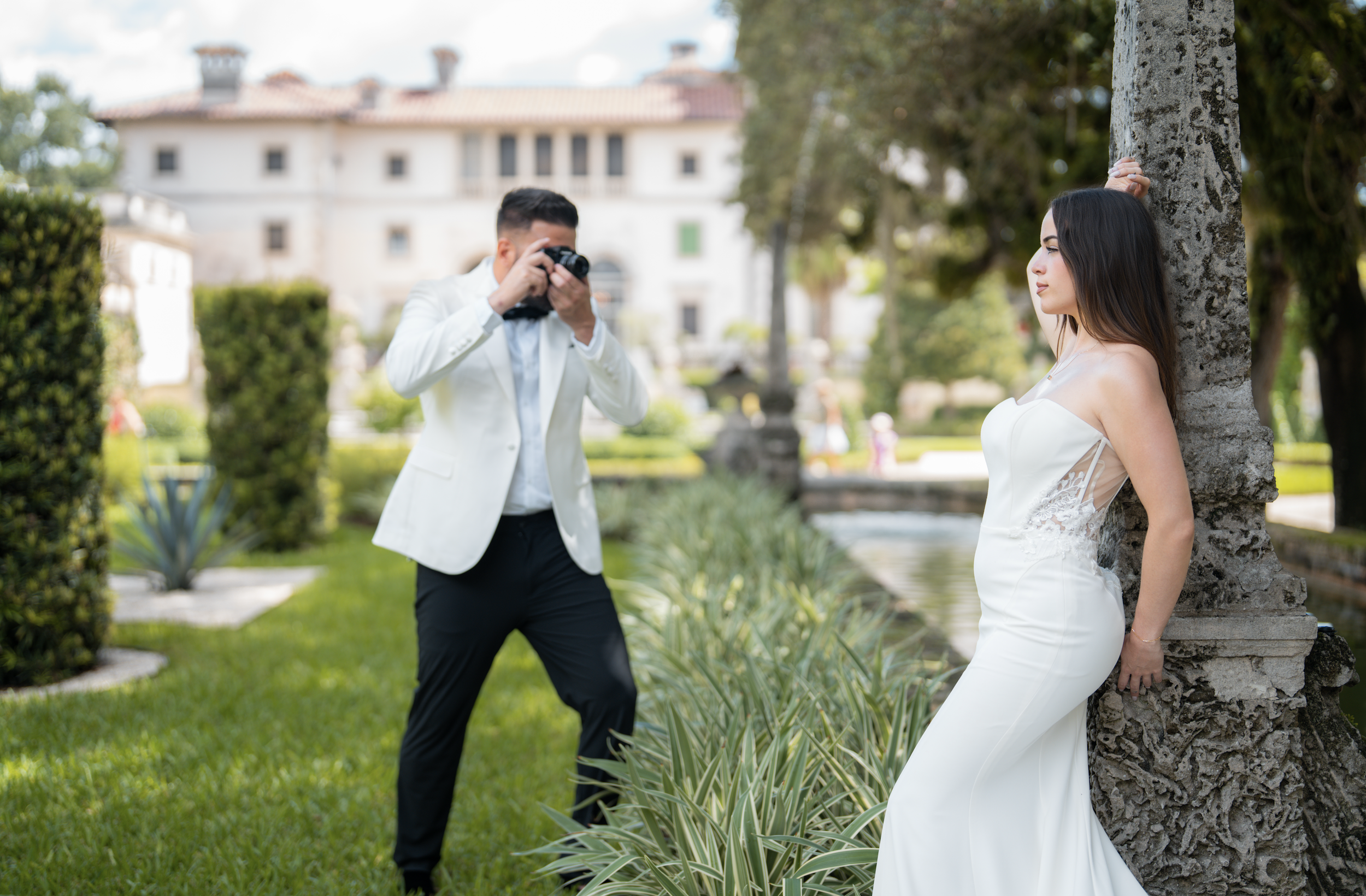 A woman in a white wedding dress leaning against a tree, while a man in a white blazer and black pants takes her photo with a camera in a garden setting.