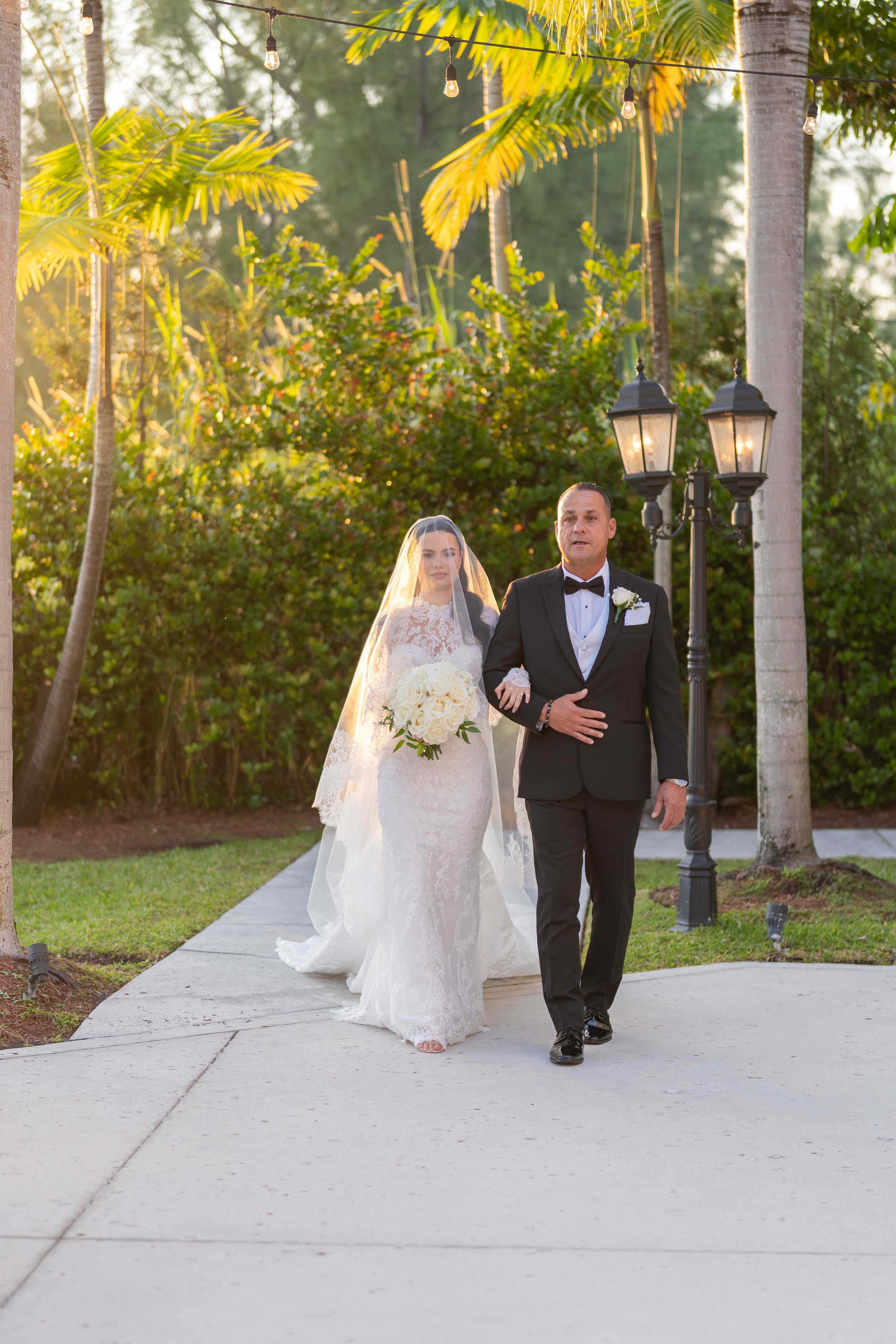 Bride walking down the aisle with her father during a wedding ceremony outdoors, surrounded by greenery and palm trees, with warm sunlight and street lamps in the background.
