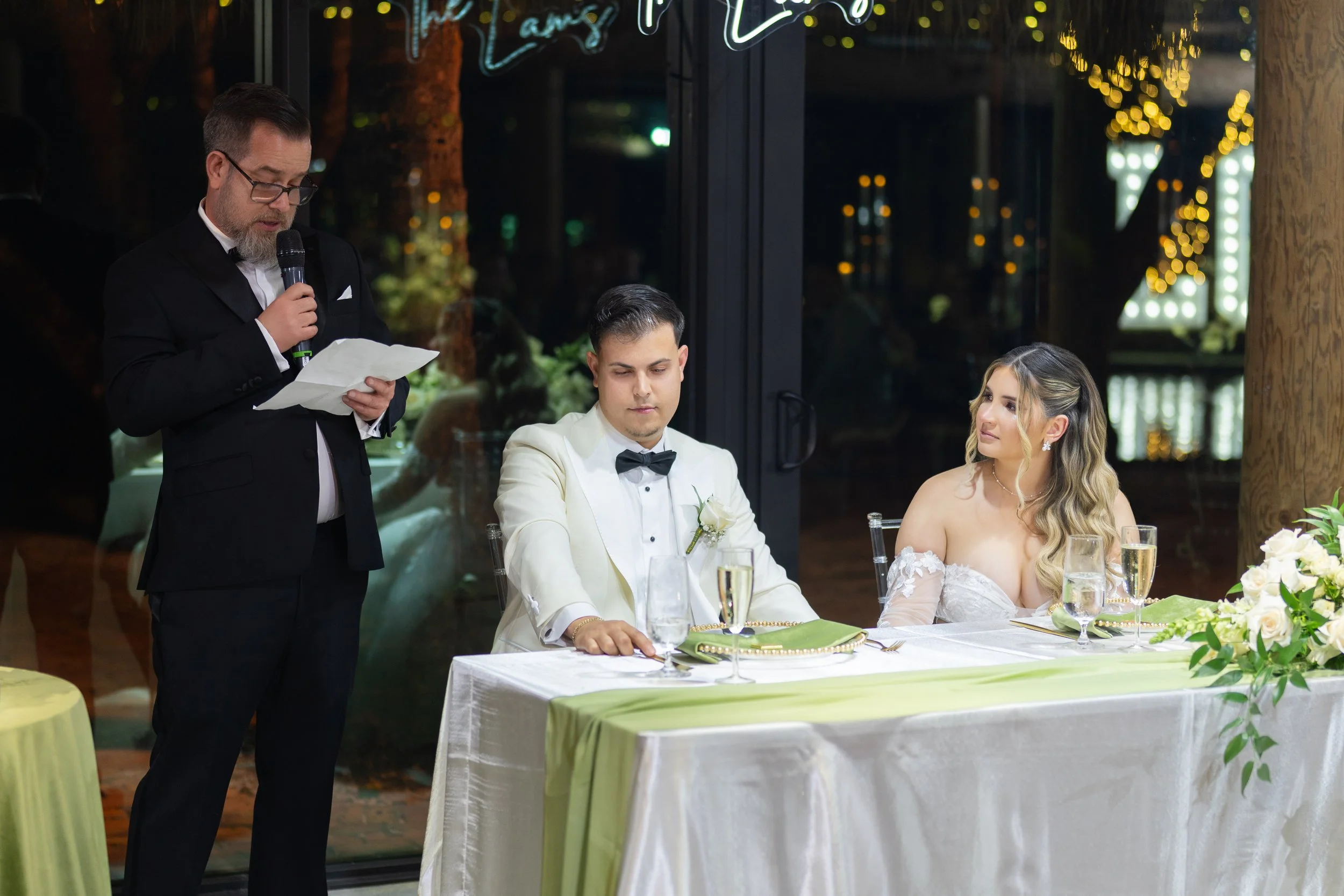 A wedding reception with a bride and groom sitting at a decorated table, the groom in a white tuxedo, and a man giving a speech with a microphone.