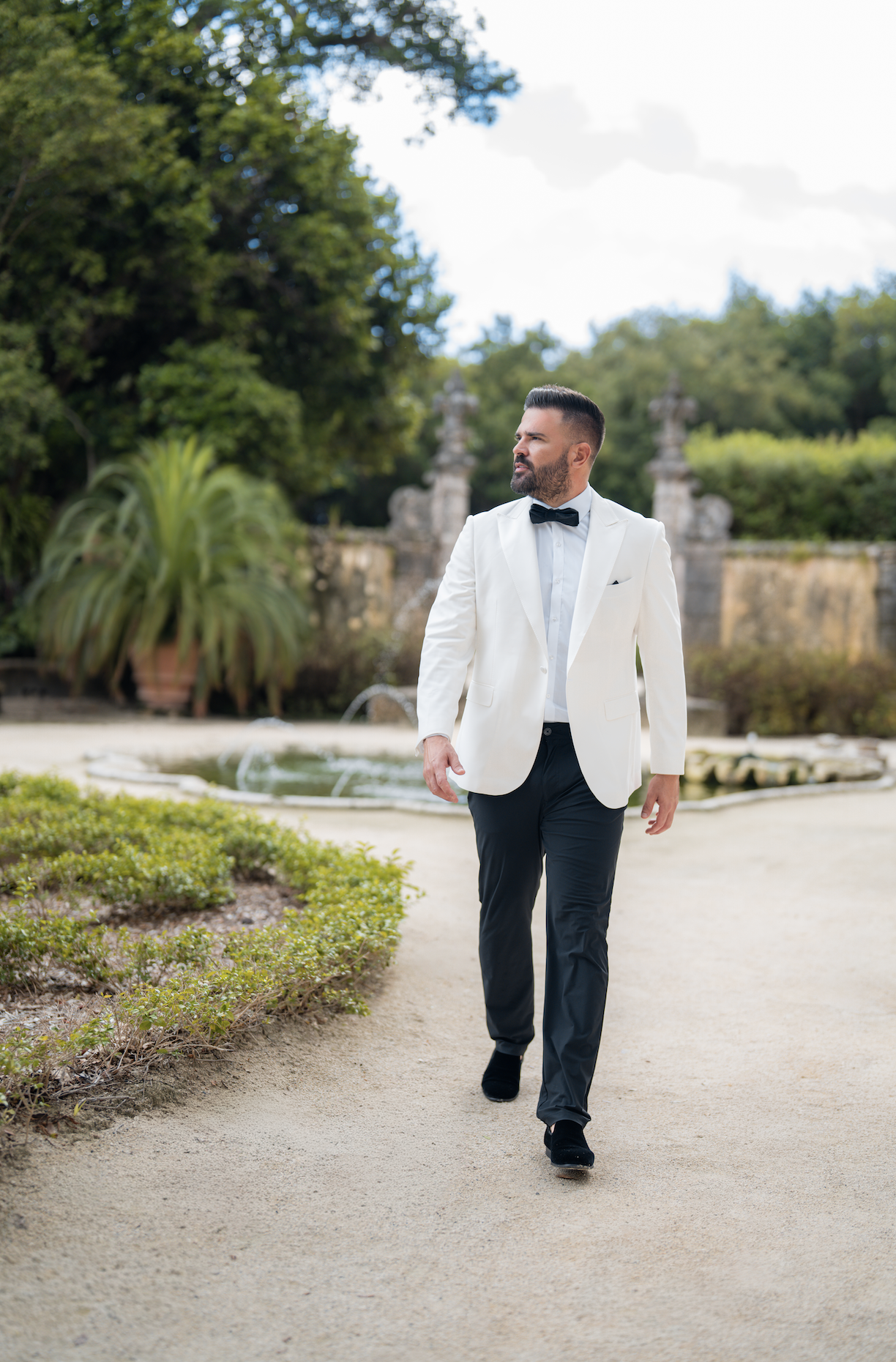 A man in a white tuxedo jacket, black pants, black shoes, and a black bow tie walking outdoors on a light-colored dirt path.