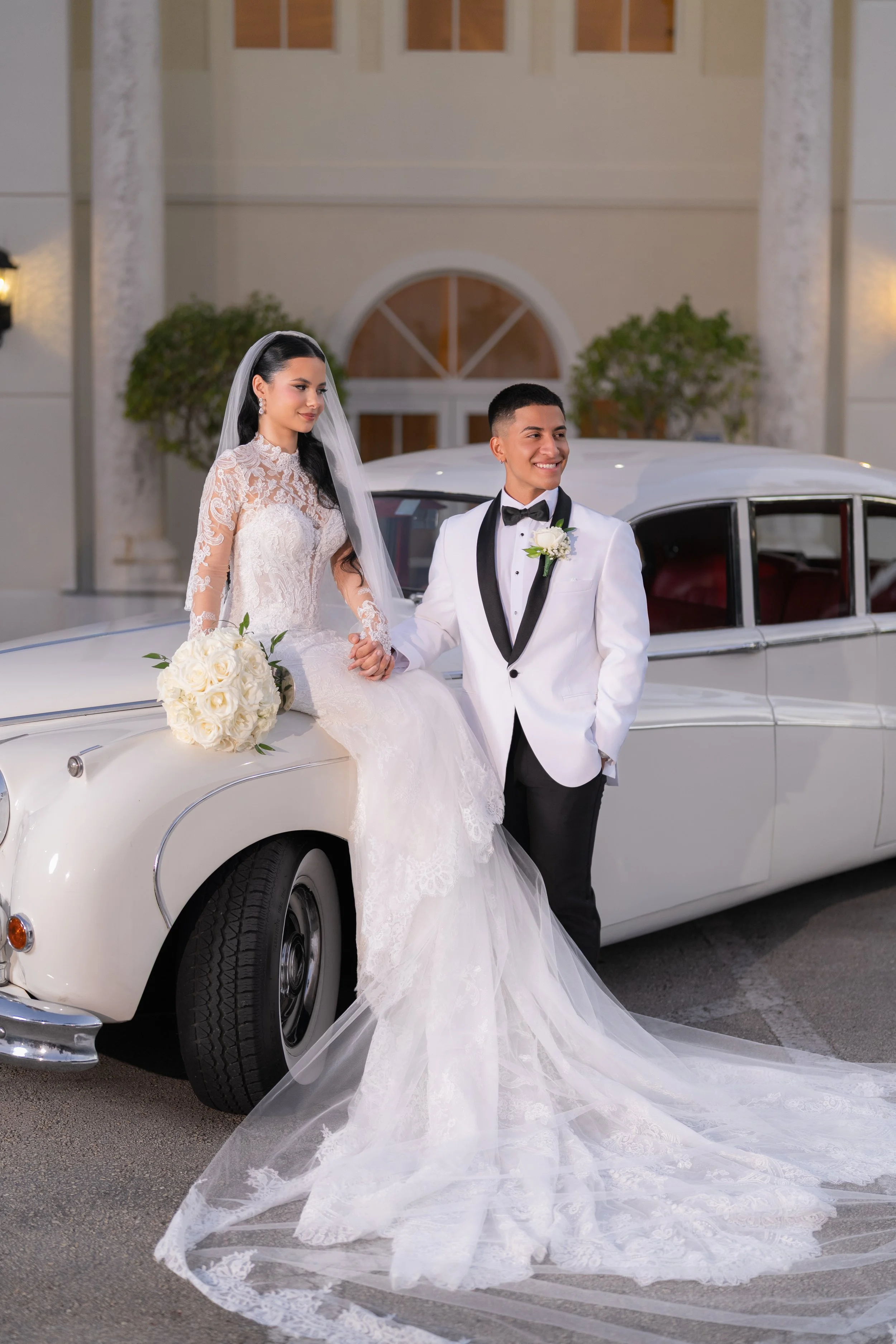 A bride and groom in wedding attire posing next to a vintage white car in front of a building.