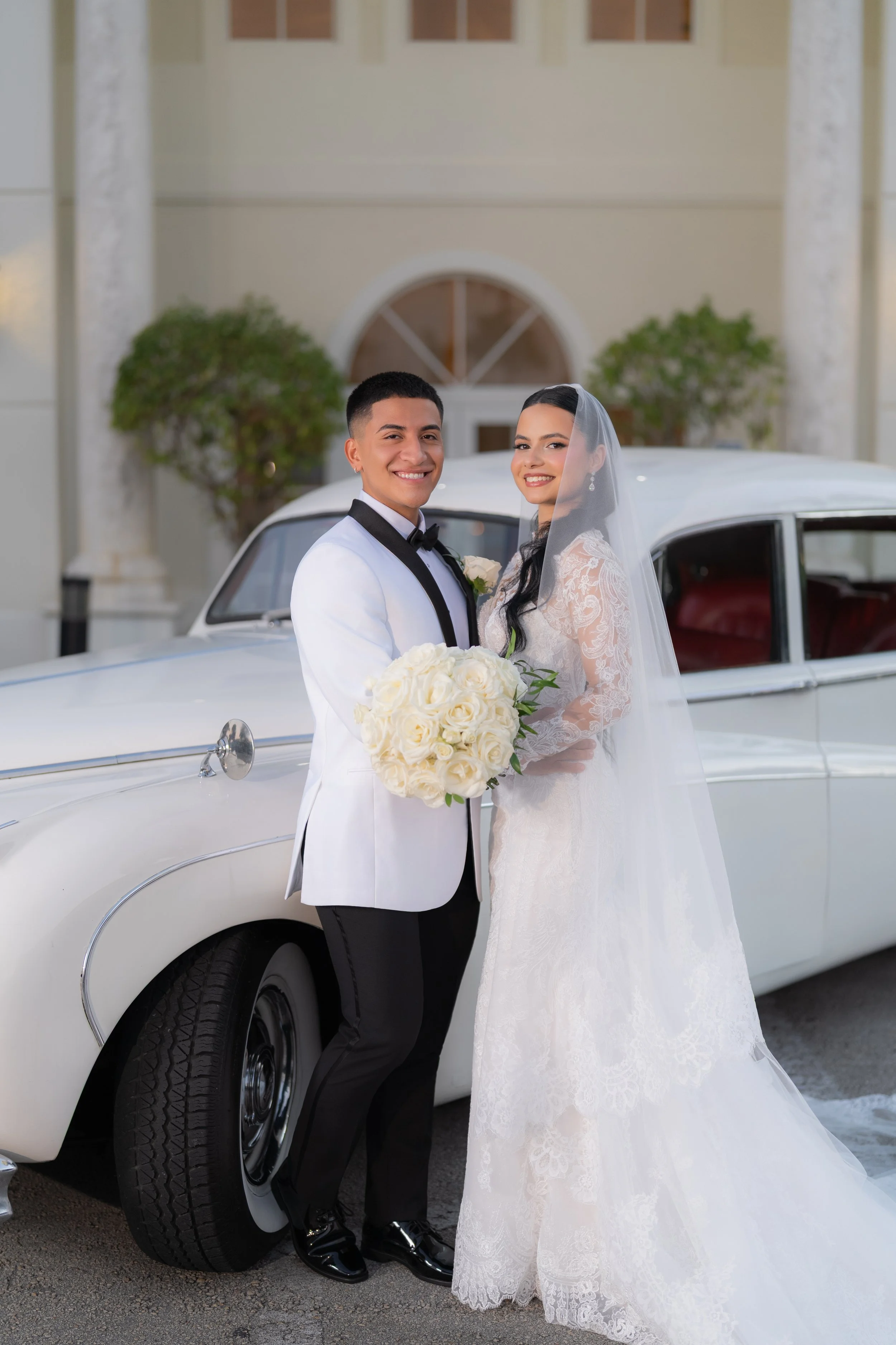 A newlywed couple in wedding attire standing next to a vintage white car, with the groom holding a bouquet of cream-colored roses, outside a building with columns and greenery.