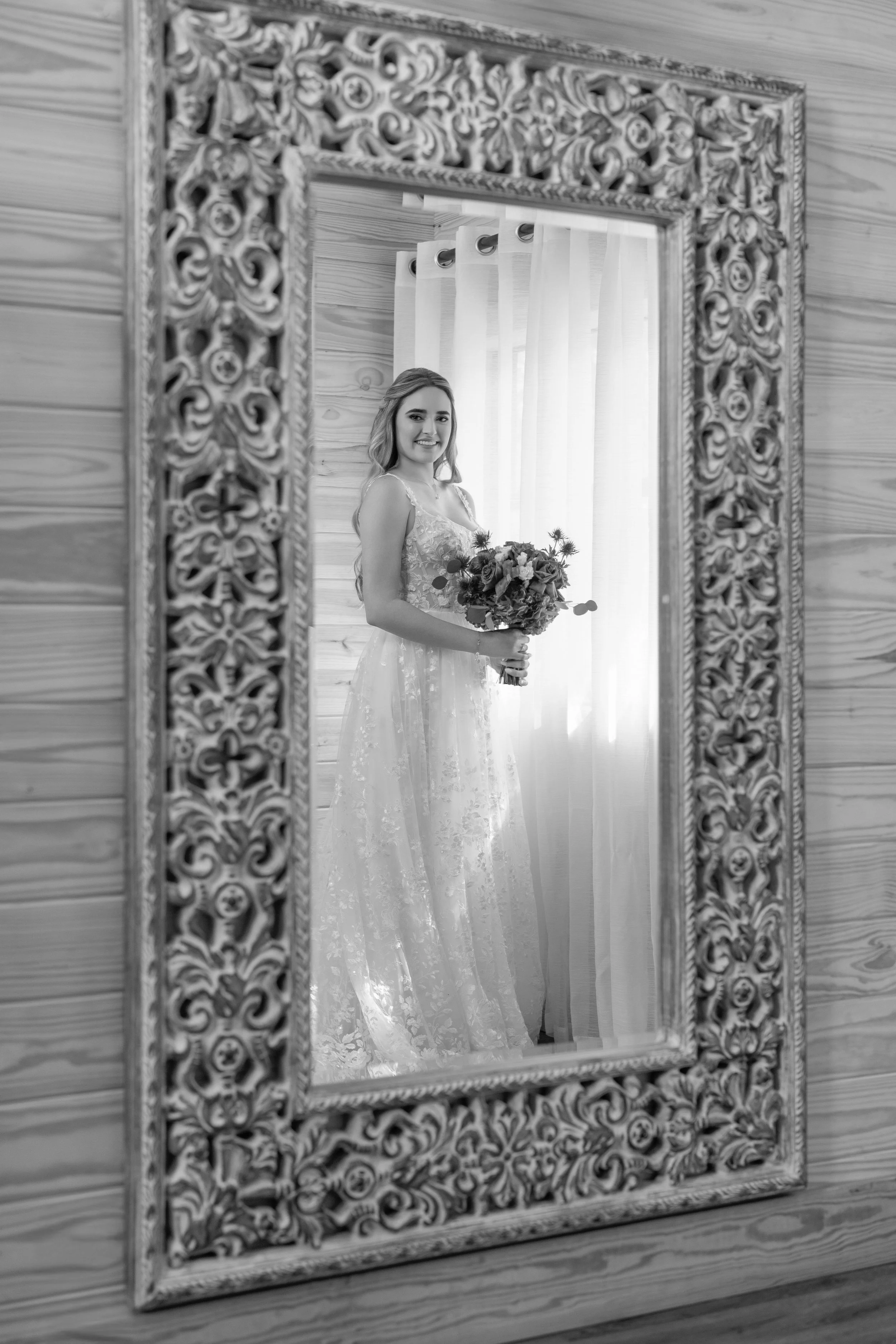 A woman in a wedding dress holding a bouquet of flowers is reflected in a decorative mirror, with a window and curtains in the background.