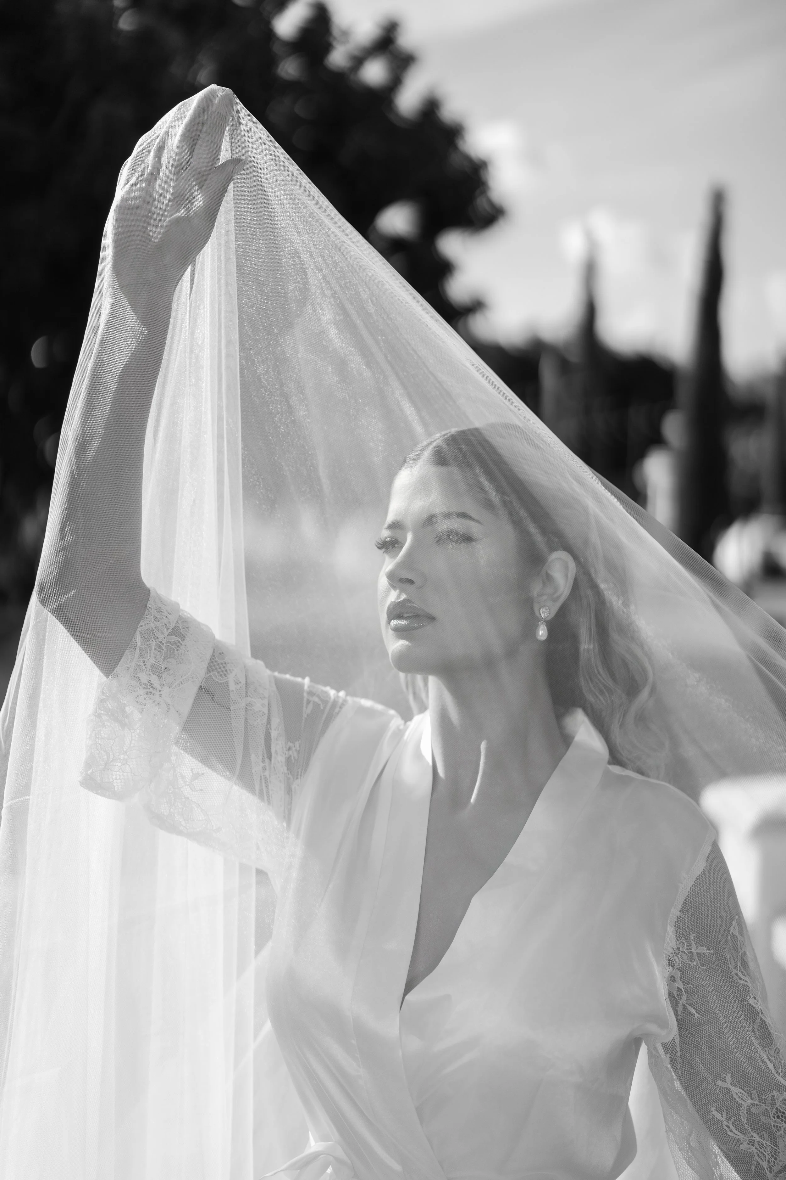 Black and white photo of a woman with wavy hair wearing earrings and a satin-like robe, lifting a sheer veil over her head outdoors.
