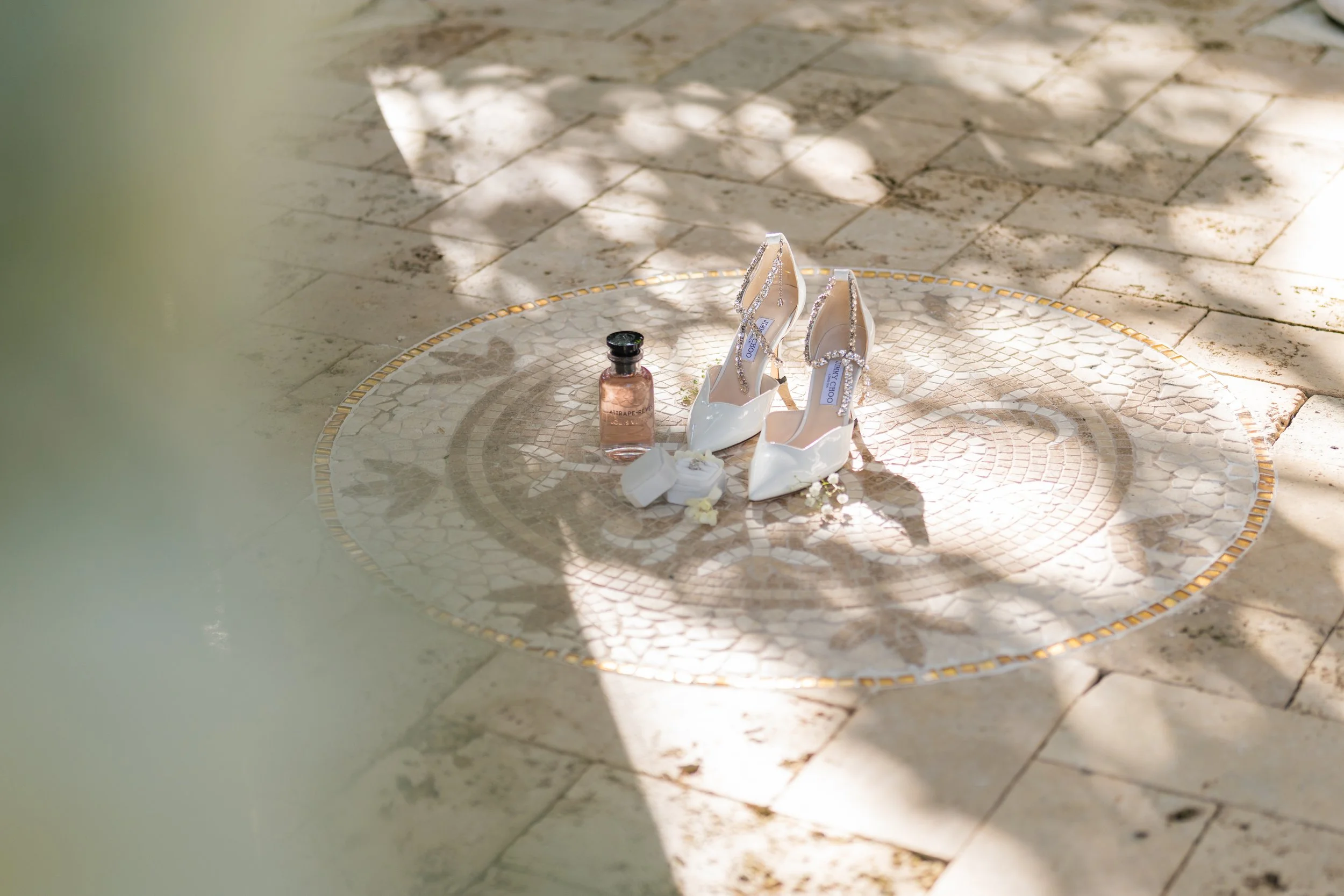 A pair of white high-heeled shoes with ankle straps, a small bottle of perfume, and a ring are arranged on a decorative mosaic circular mat on a stone floor, illuminated by sunlight and surrounded by shadows.