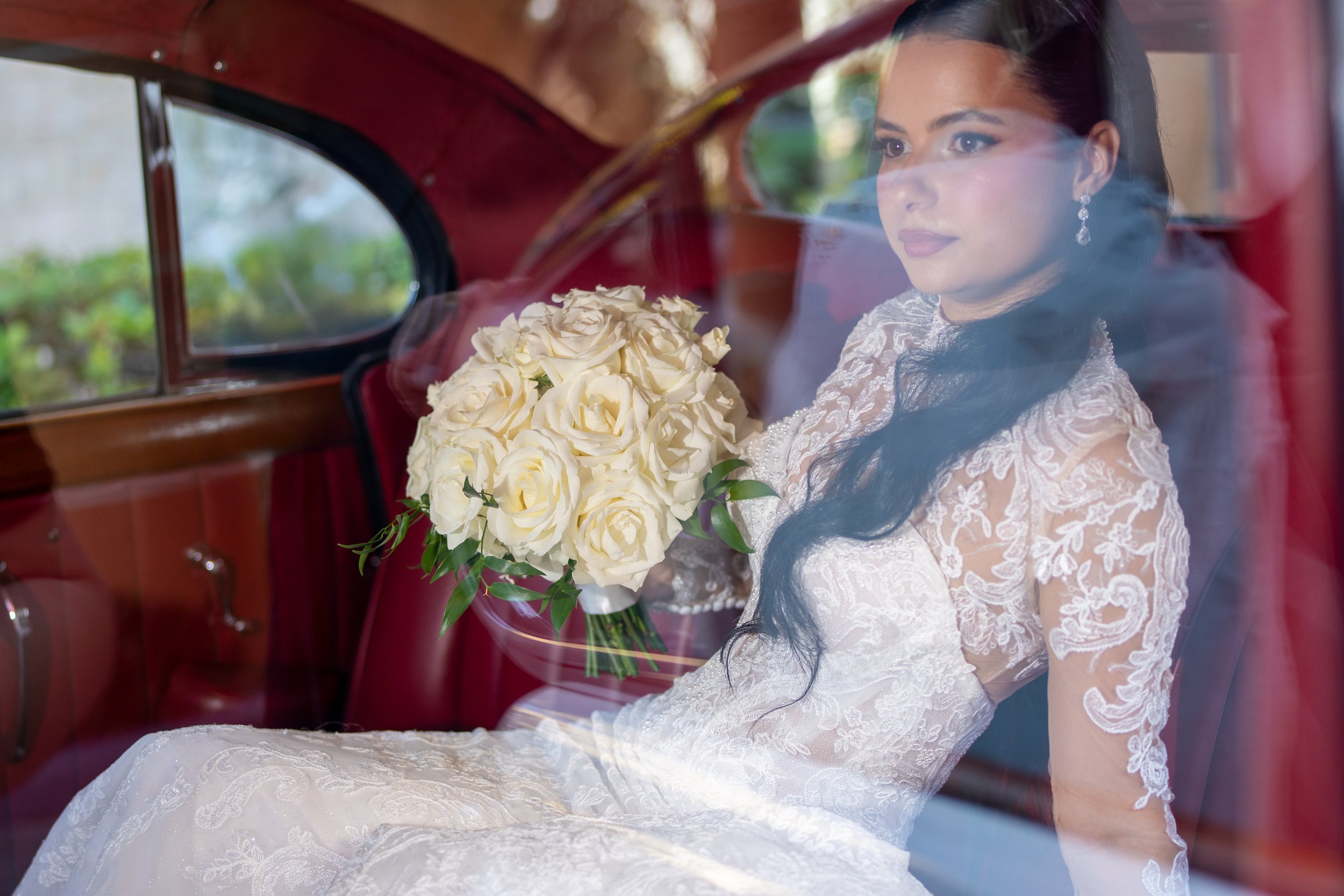 A bride with long dark hair wearing a lace wedding dress sitting inside a vintage car, holding a bouquet of white roses, seen through the car window.