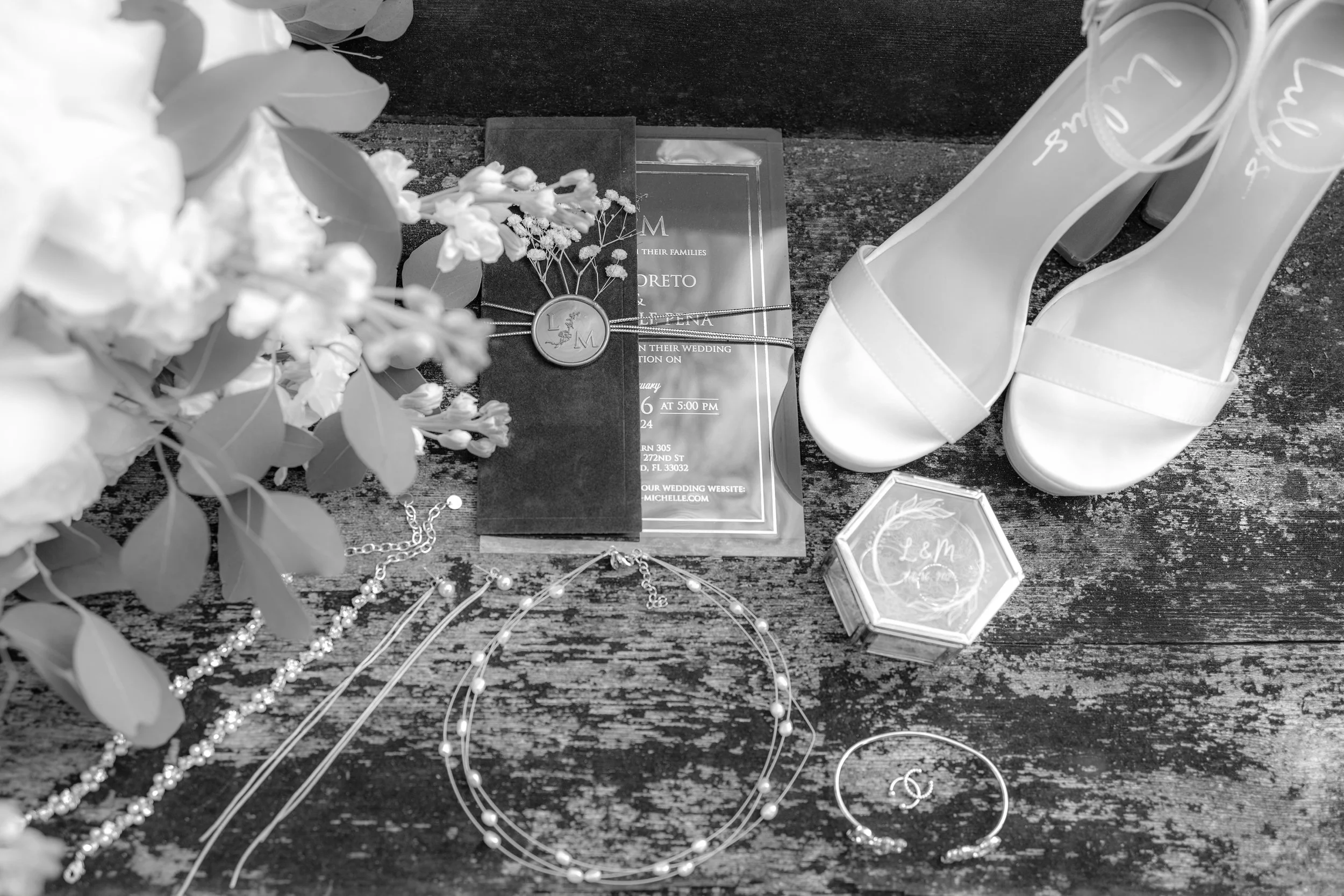 Wedding accessories and invitation on a wooden surface, including white high-heeled shoes, jewelry, a wedding invitation, a small box, and a floral bouquet.