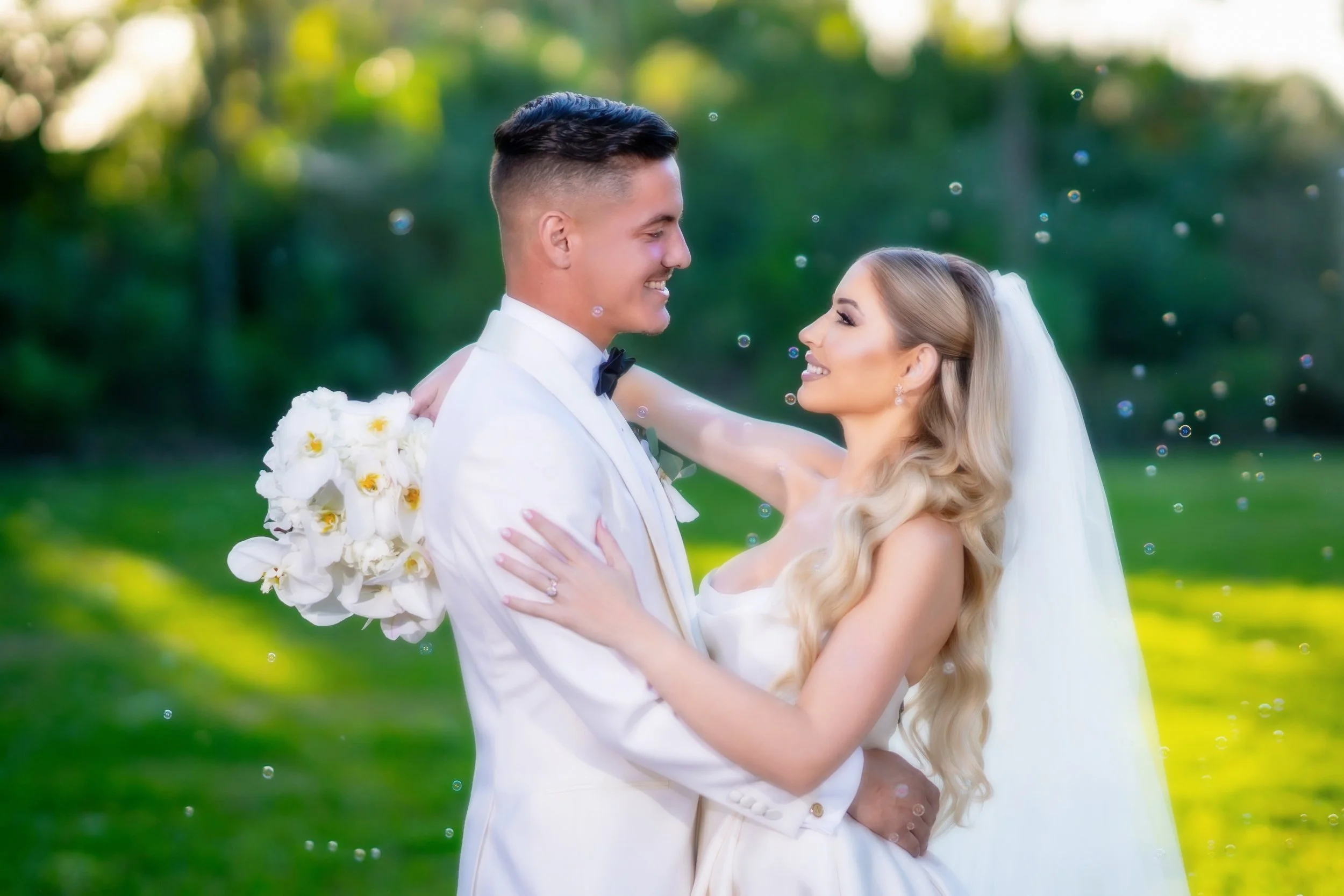A newlywed couple smiling and looking into each other's eyes outdoors with greenery in the background. The groom is holding a bouquet of white flowers behind his back and wearing a white tuxedo with a black bow tie. The bride has long blonde hair, ve