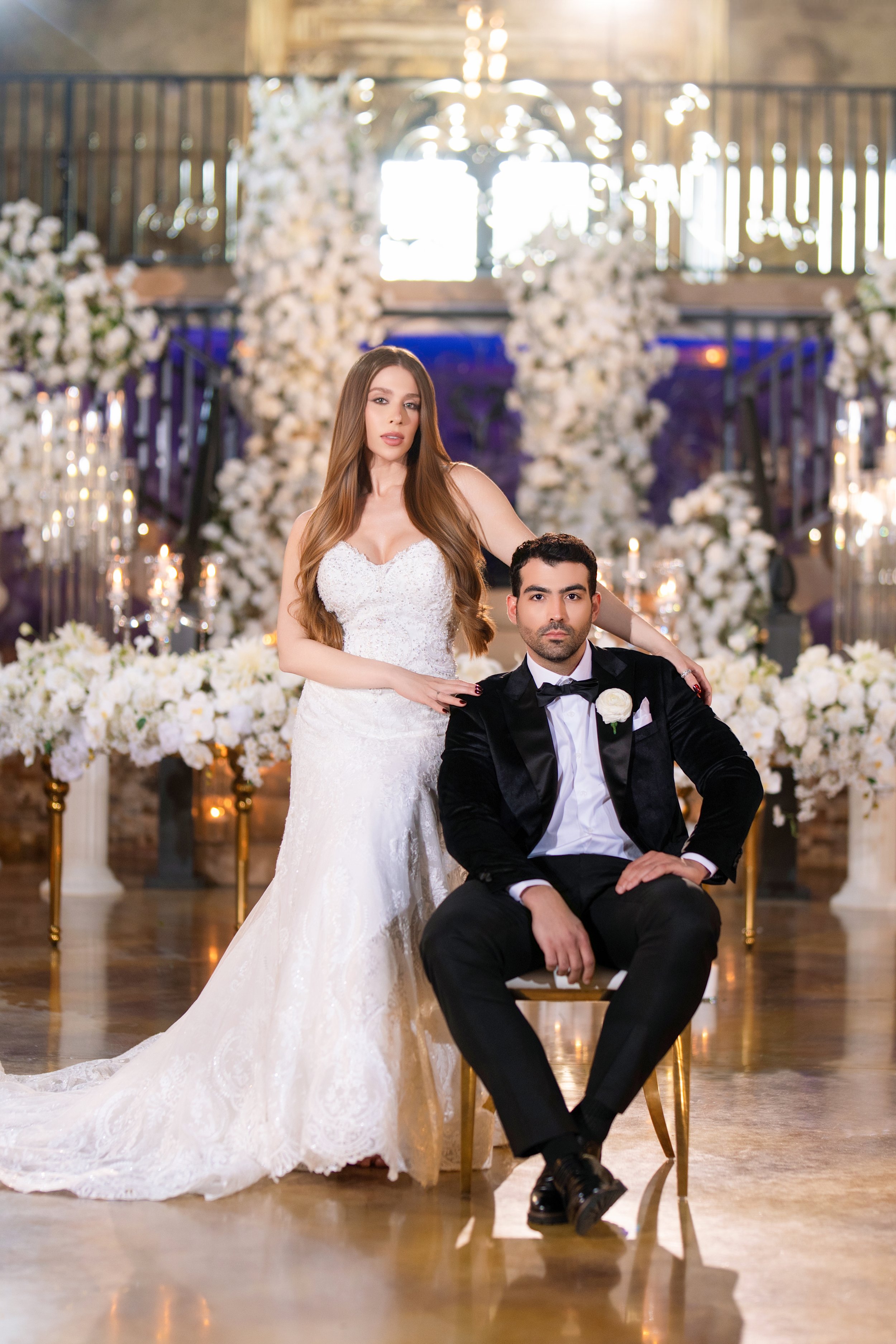 A bride and groom poses for a wedding photo in a decorated venue with floral arrangements and candles.