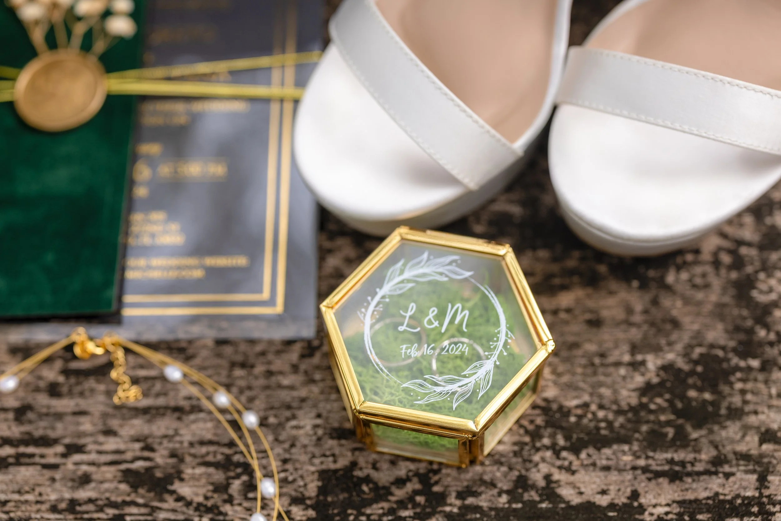 Wedding jewelry, white shoes, and a personalized glass box with the initials 'L & M' and the date 'Feb 16, 2024' on a wooden surface.
