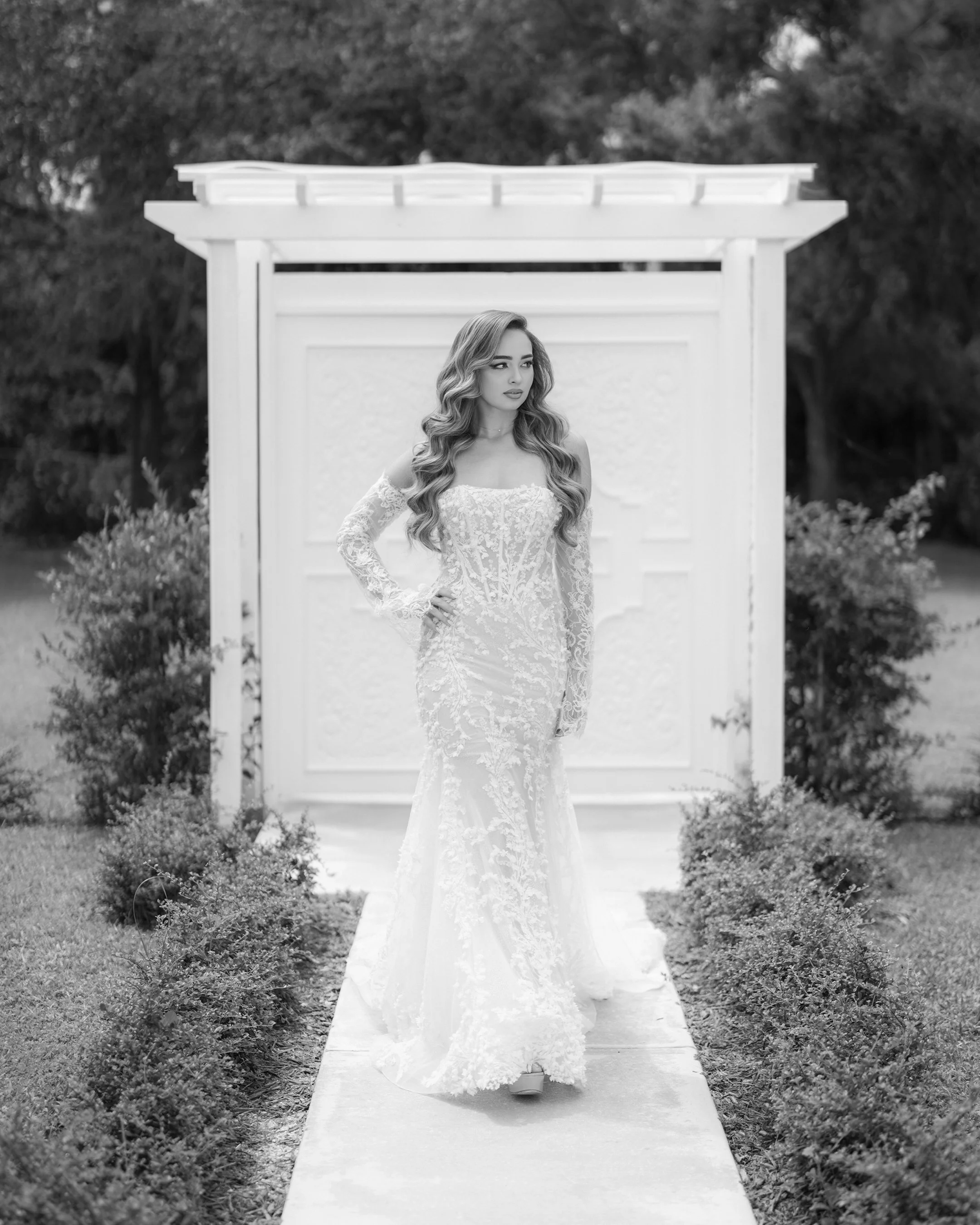 A woman in a lace wedding dress walking down a garden path in front of a white decorative gate.