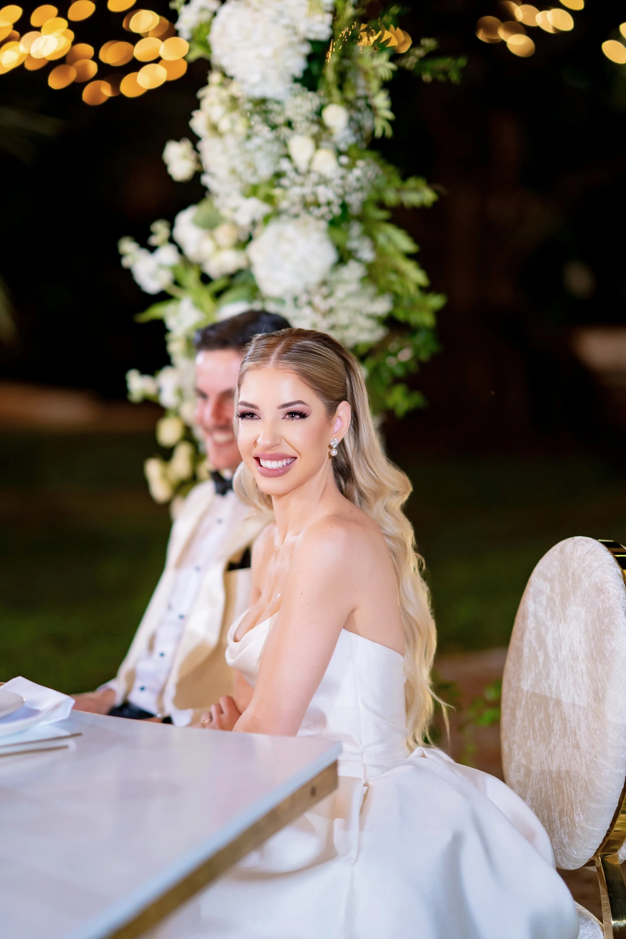 A smiling bride in a wedding dress sitting at a table during a wedding reception, with a groom beside her and a floral backdrop behind them.