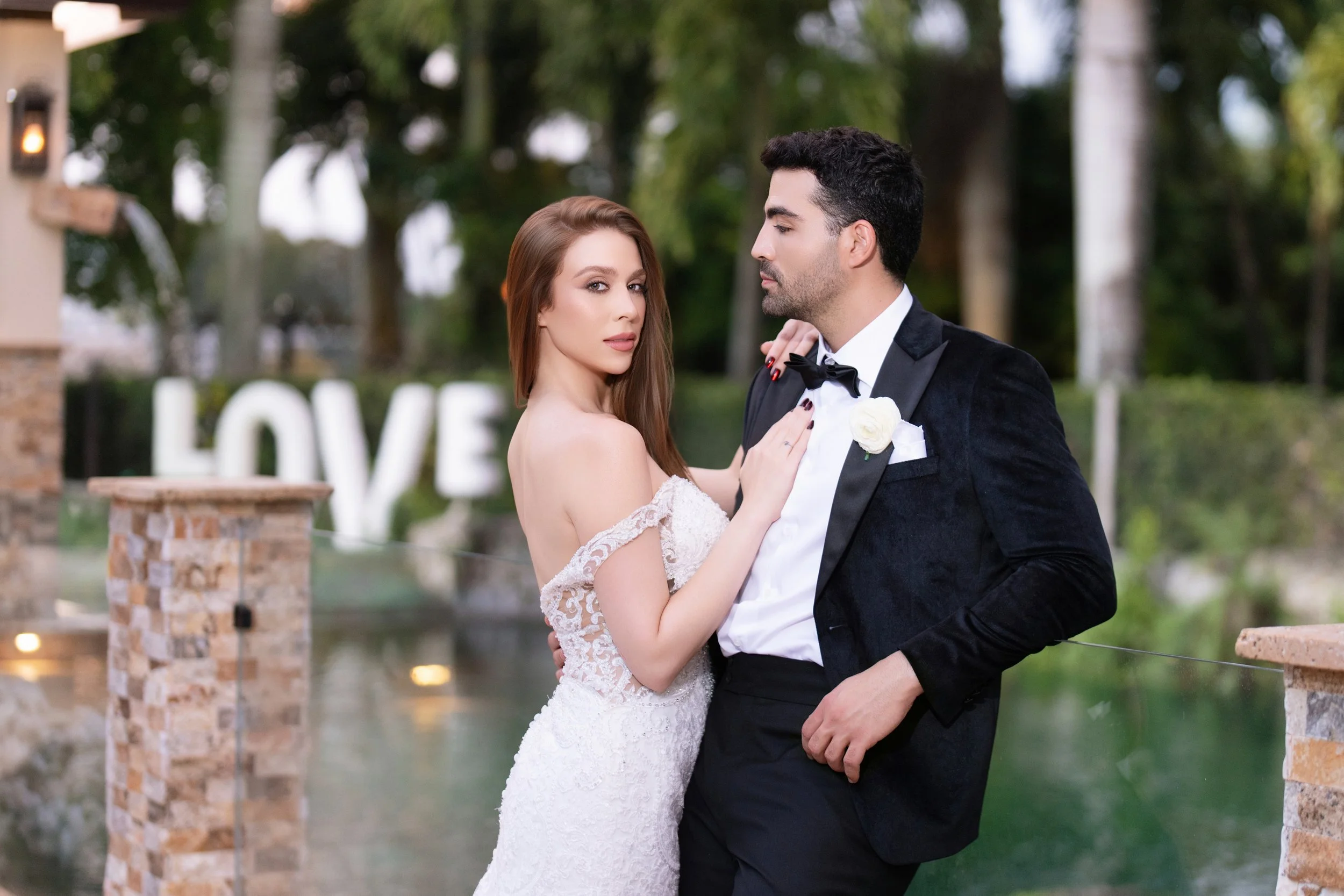 A bride and groom in wedding attire by a water feature with a large illuminated 'LOVE' sign in the background.