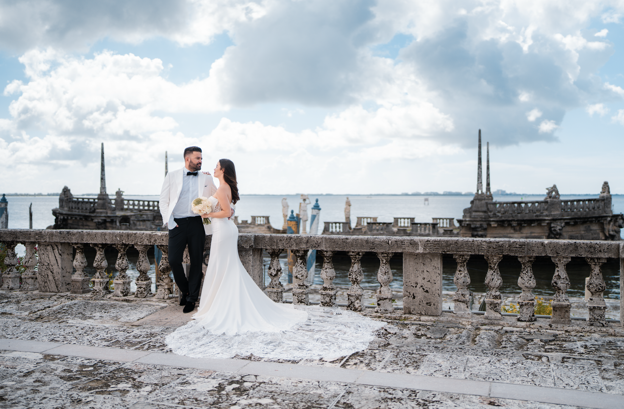 Bride and groom standing on a stone terrace by a body of water, with a cloudy sky in the background, holding a bouquet, dressed in wedding attire.