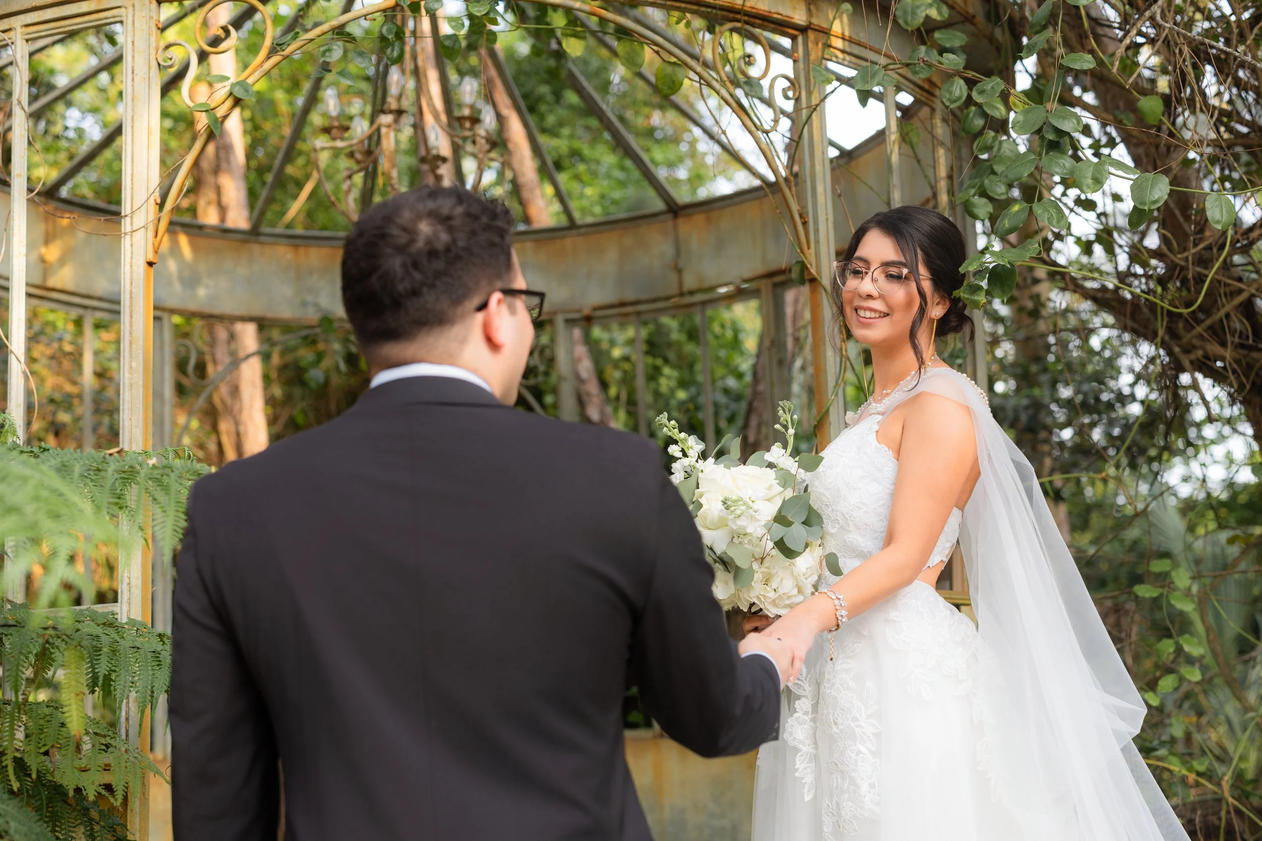 A bride and groom holding hands during their wedding ceremony outdoors in a garden, with the bride in a white wedding dress holding a bouquet of white flowers, and the groom in a black suit.
