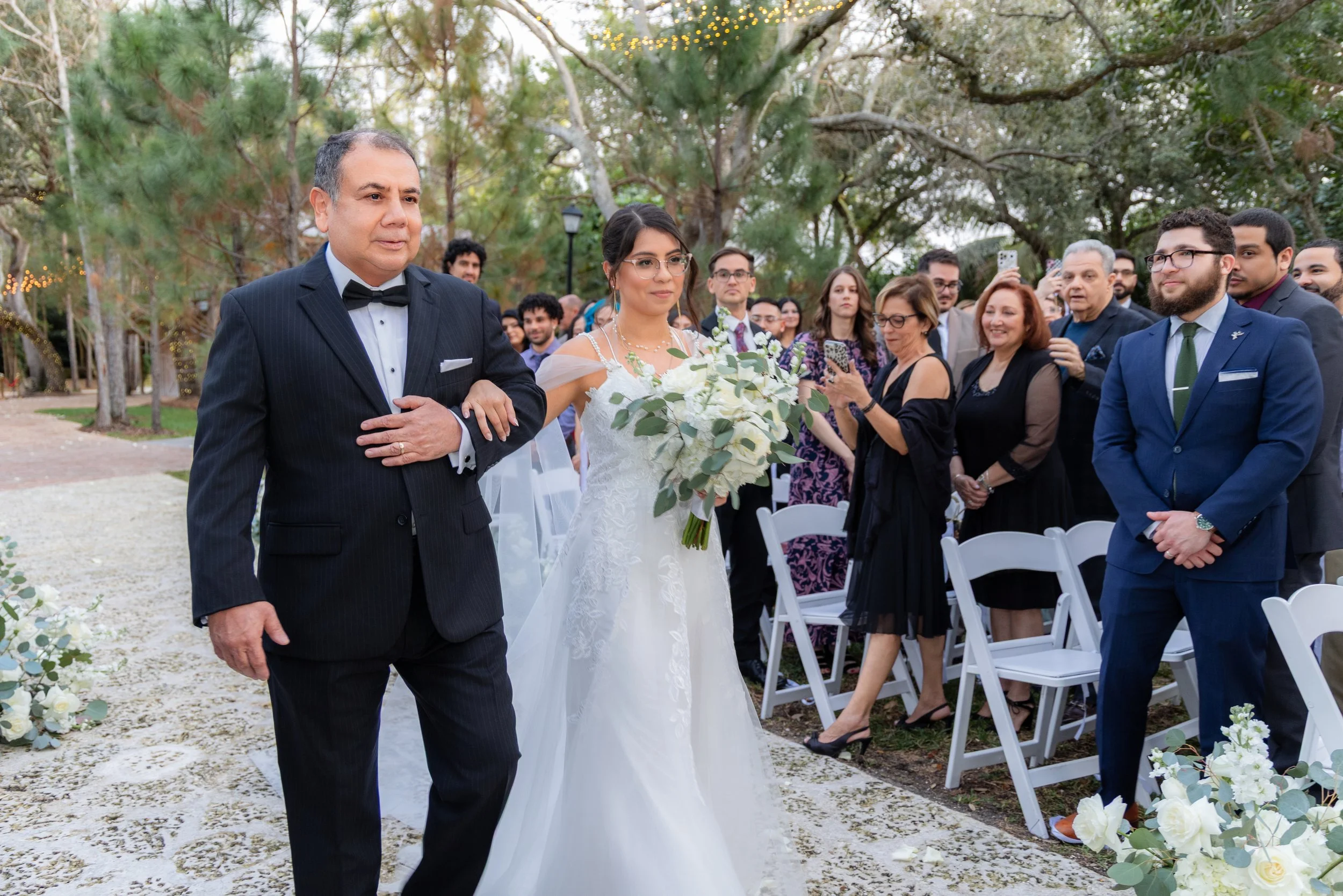 A bride walking down the aisle with her father, holding a bouquet of white flowers and greenery at an outdoor wedding ceremony, while guests watch and smile.