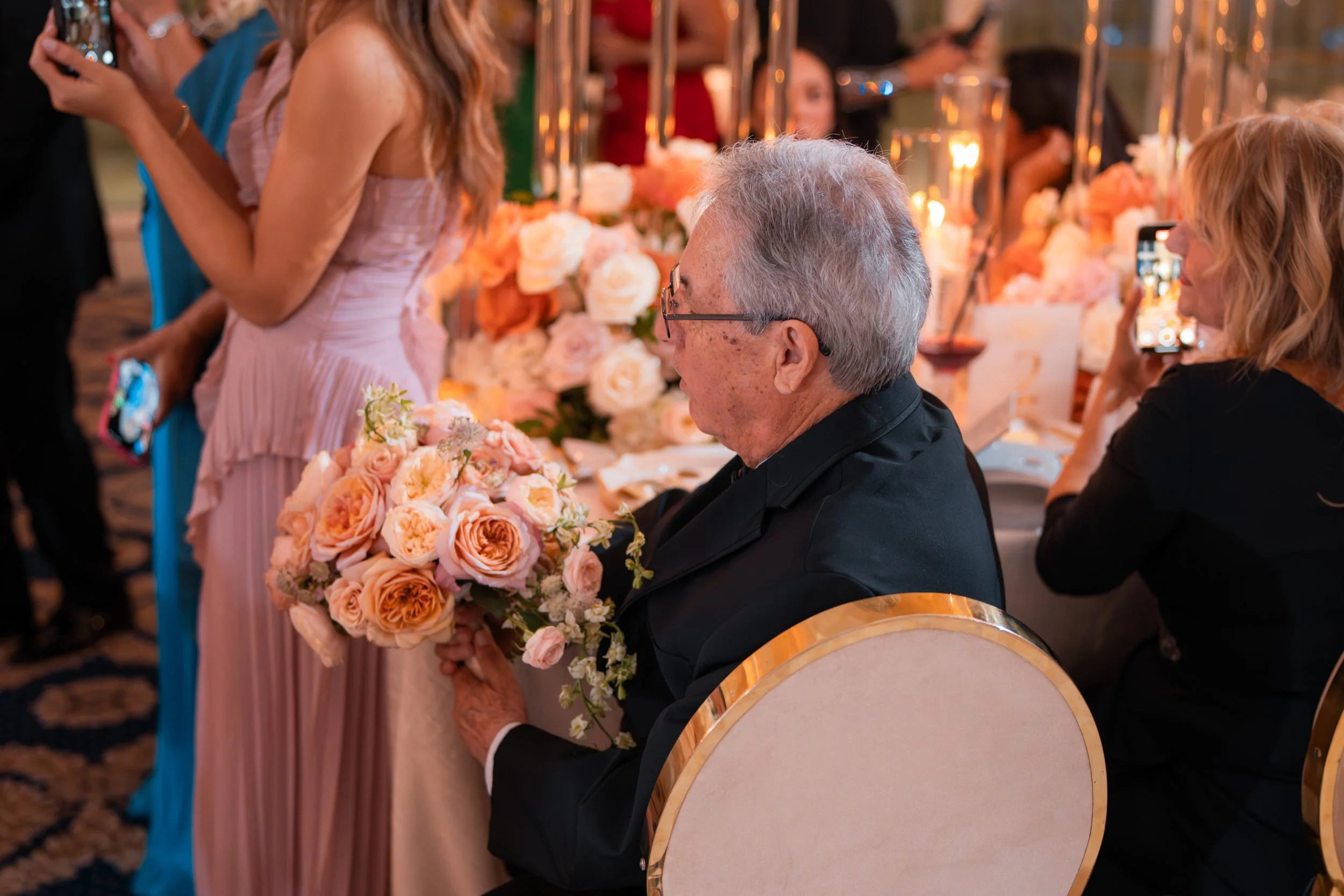 Older man sitting at a table with a bouquet of pink and peach roses, surrounded by women at a formal event with floral and candle decorations.