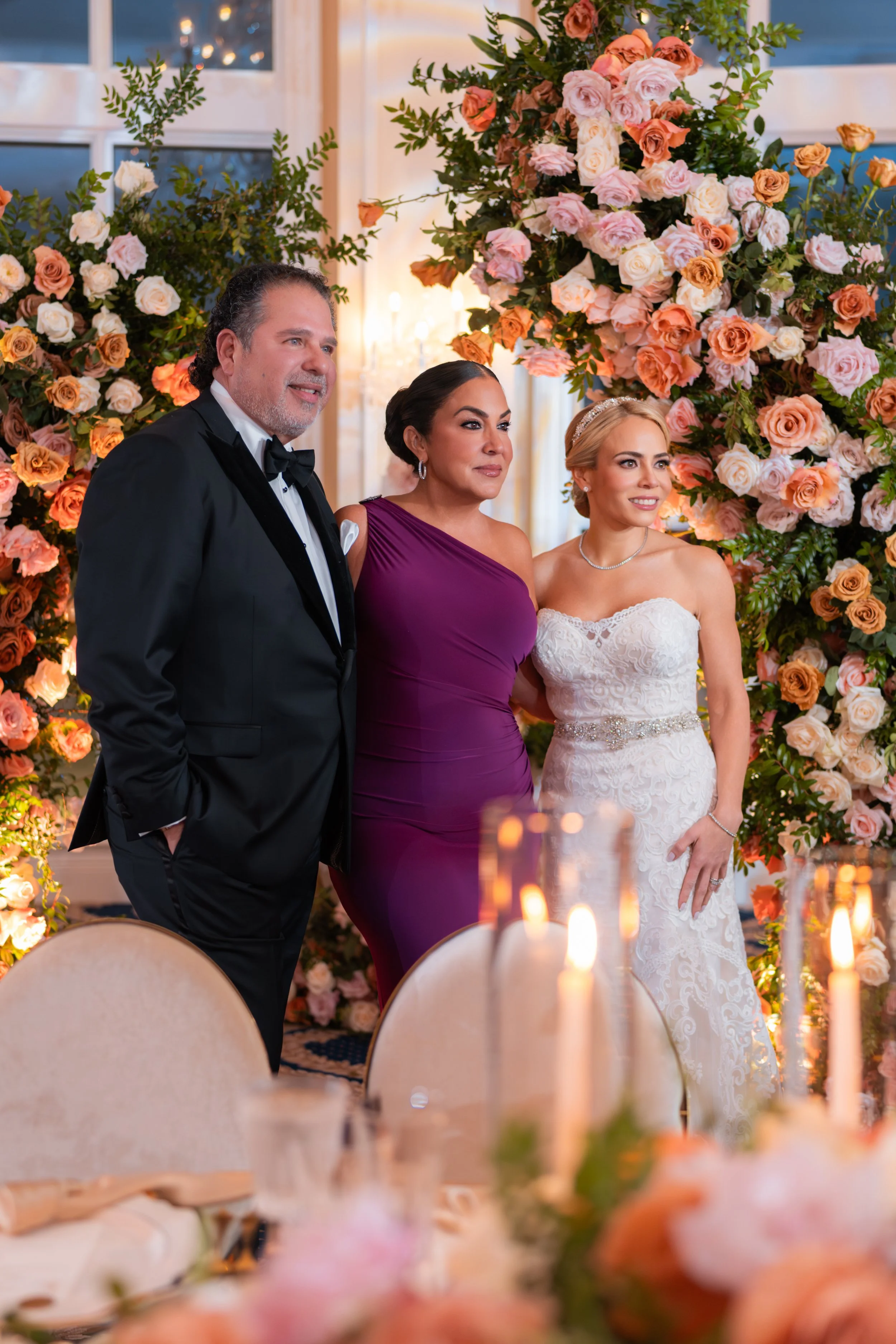 A bride in a white lace wedding gown, a man in a black tuxedo, and a woman in a purple dress pose together at a wedding reception, in front of a floral backdrop of pink, peach, and white roses.