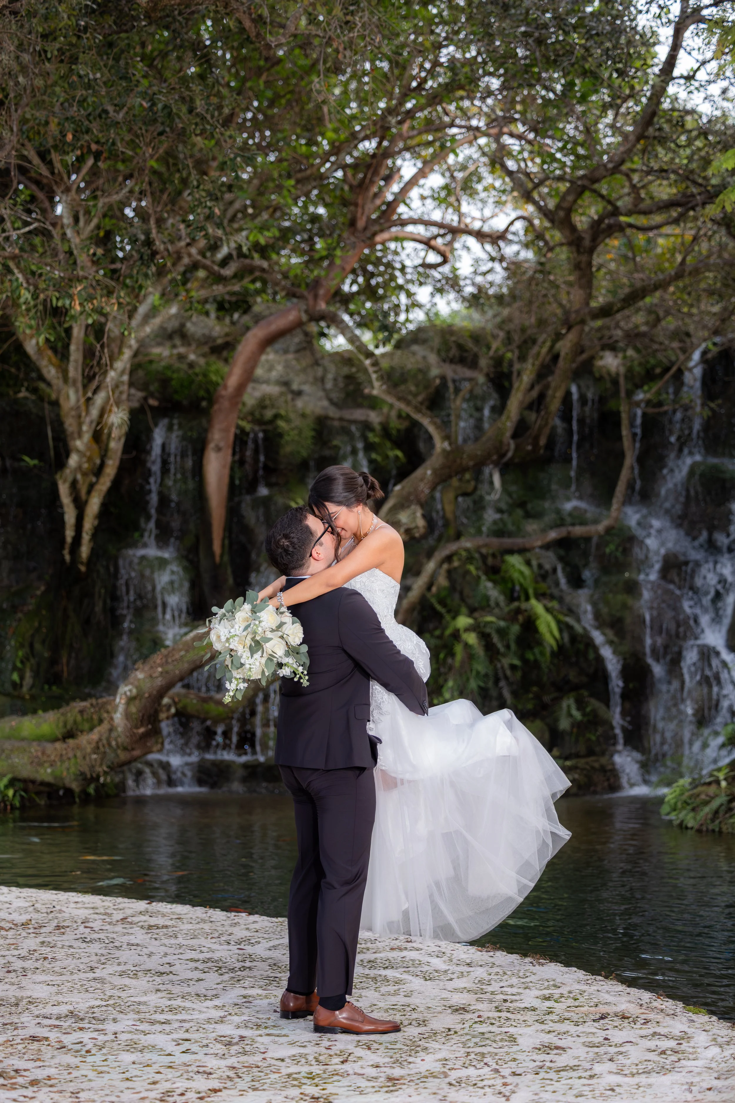 A newlywed couple in wedding attire, with the groom lifting the bride, kissing, and holding a bouquet, in front of a waterfall with trees and water in the background.