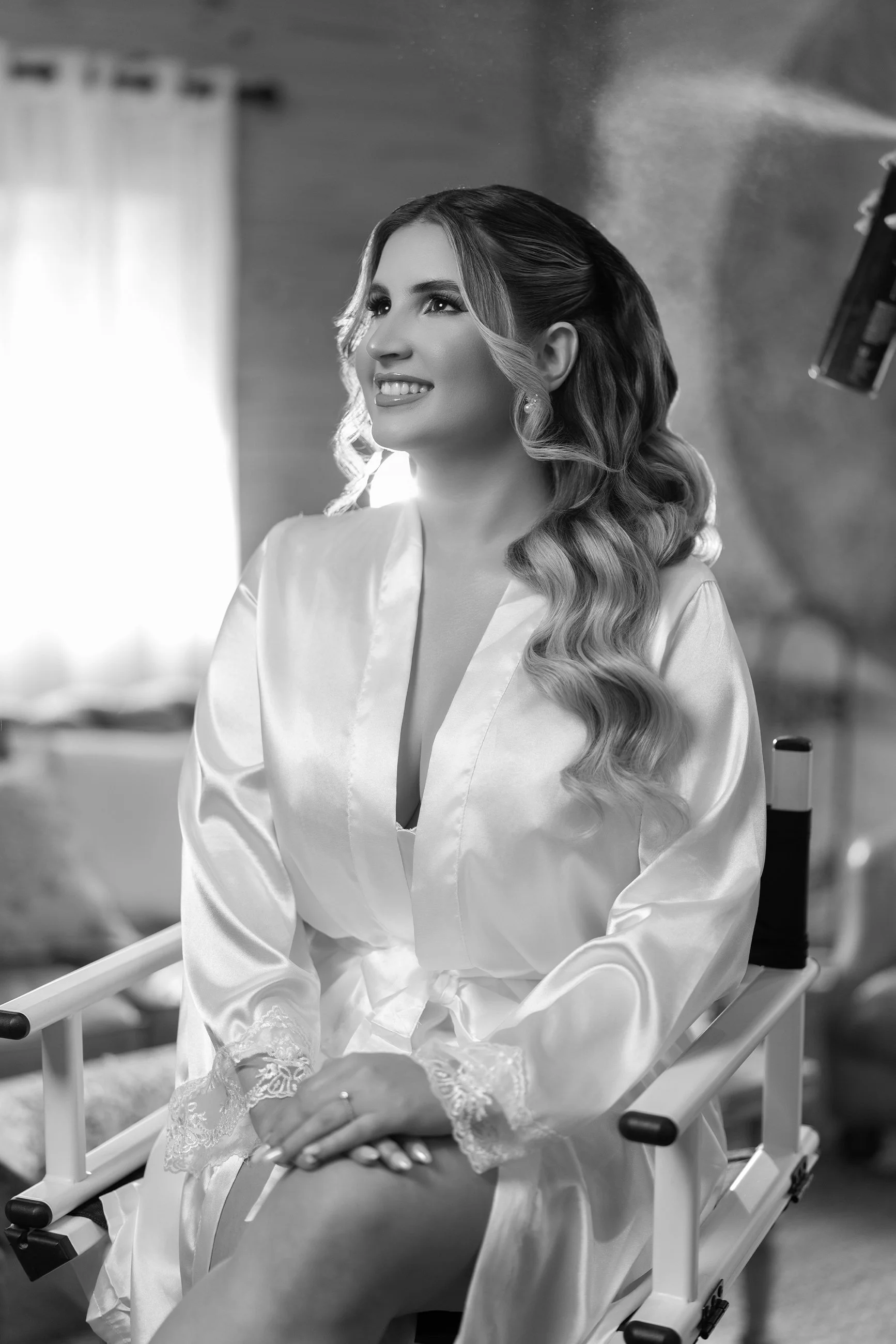 A woman sitting on a chair, dressed in a satin robe, with styled hair, smiling, in a softly lit room.