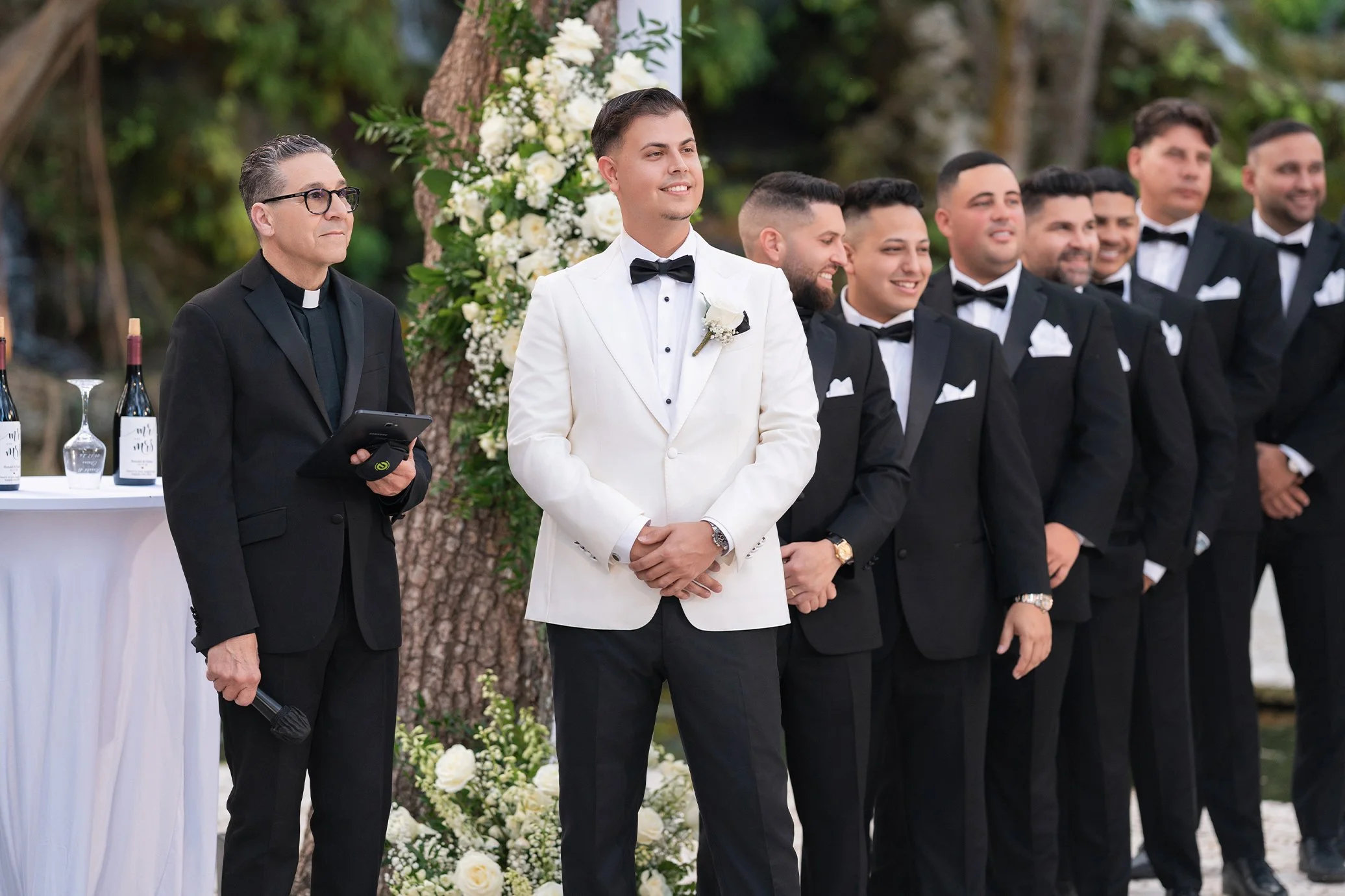 Groom in a white tuxedo standing with groomsmen in black tuxedos during outdoor wedding ceremony.