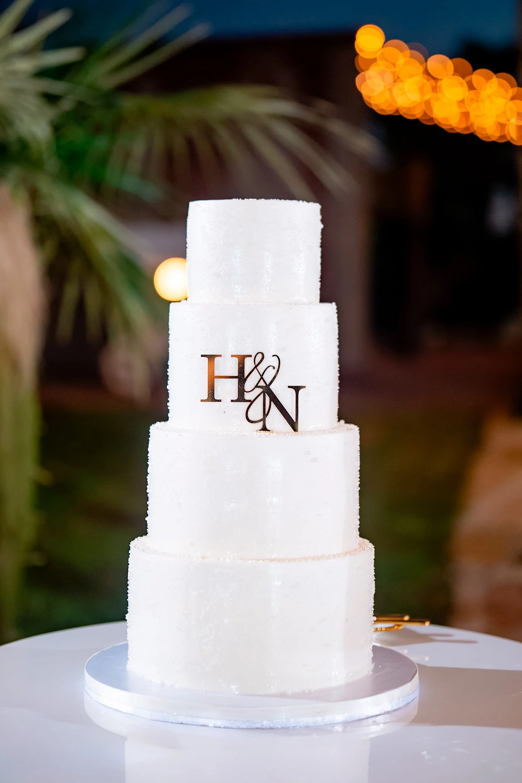 A multi-tiered white wedding cake with an illuminated base, featuring a black monogram reading 'H & N' on the second tier, placed on a white cake stand outdoors at night with blurred string lights and greenery in the background.