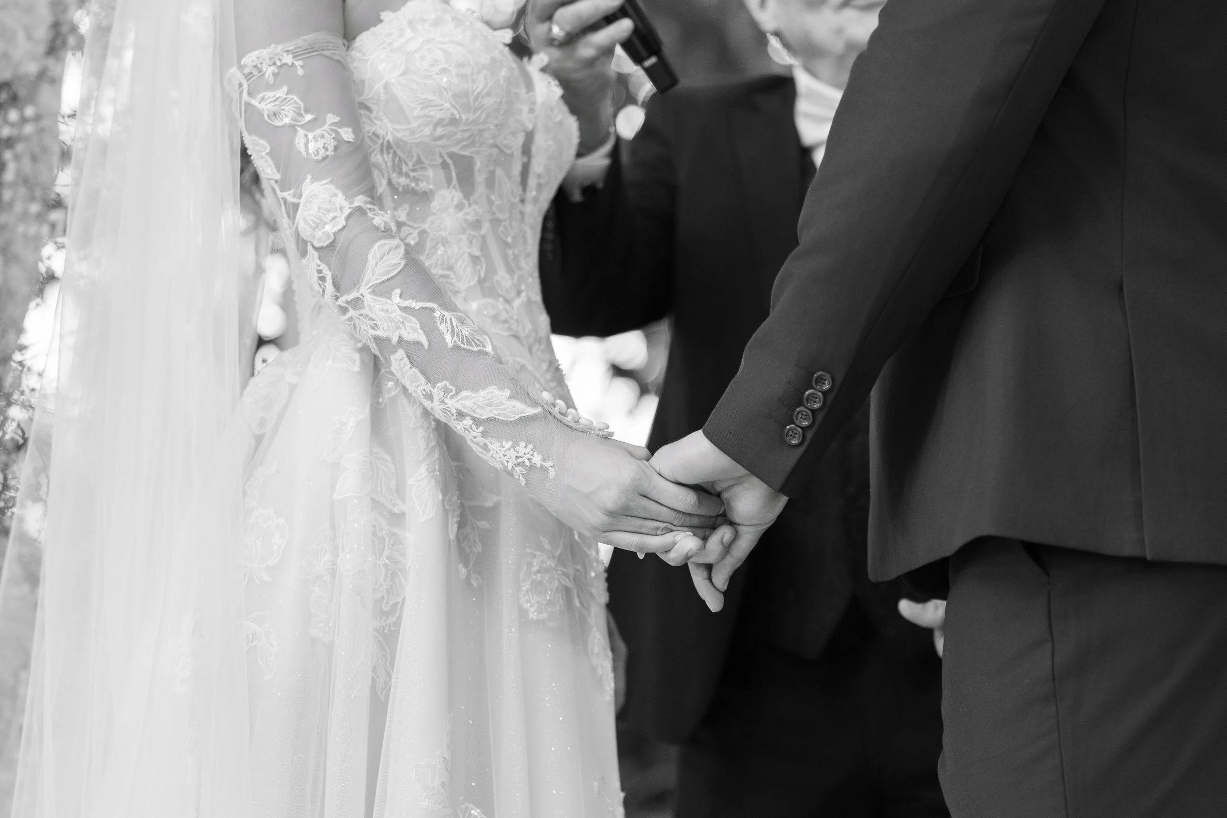 A bride and groom holding hands during a wedding ceremony, with the bride wearing a lace wedding gown and veil, and the groom in a dark suit.