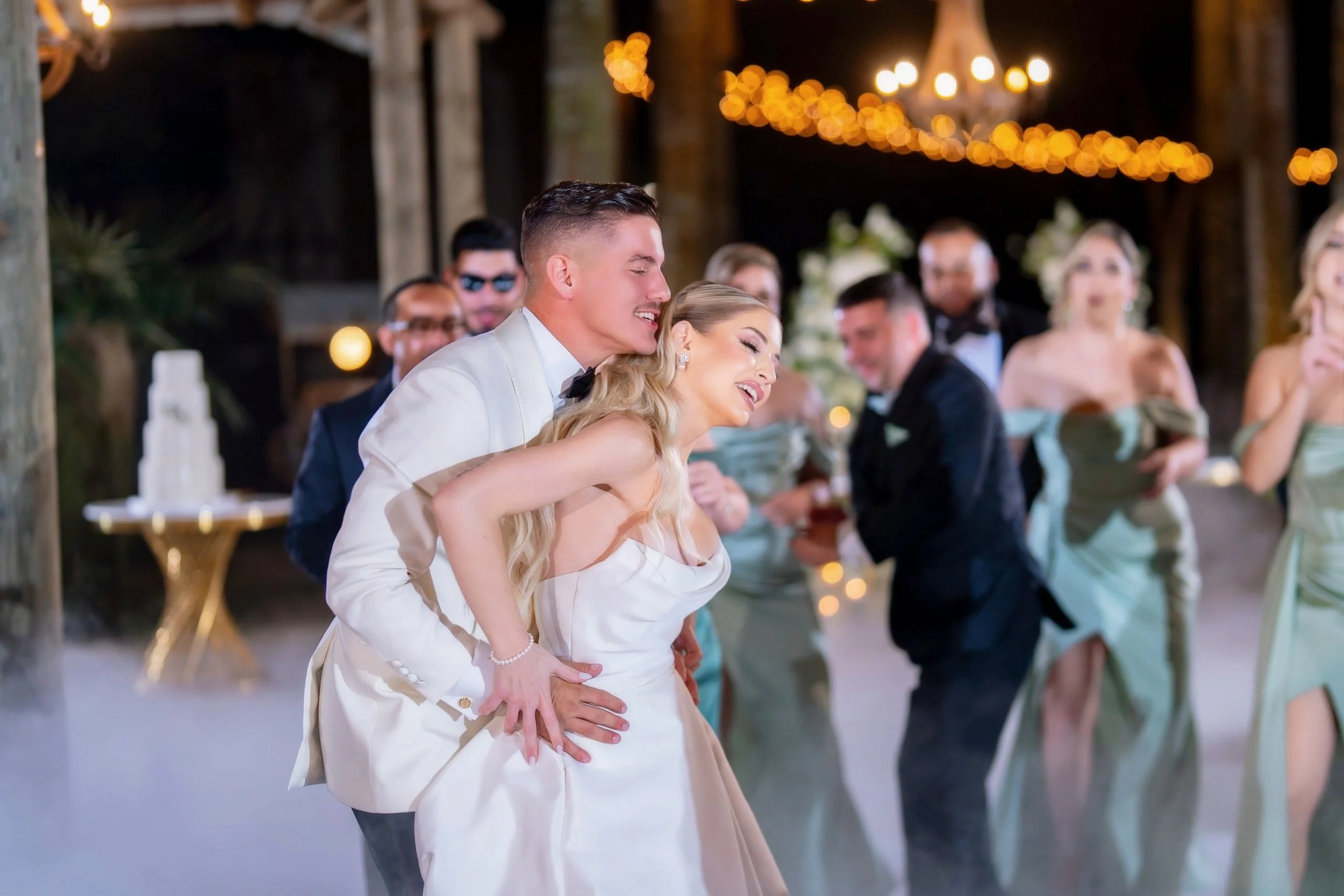 A bride and groom are dancing closely at a wedding reception with happy guests in the background, decorated with warm lights and a cake.