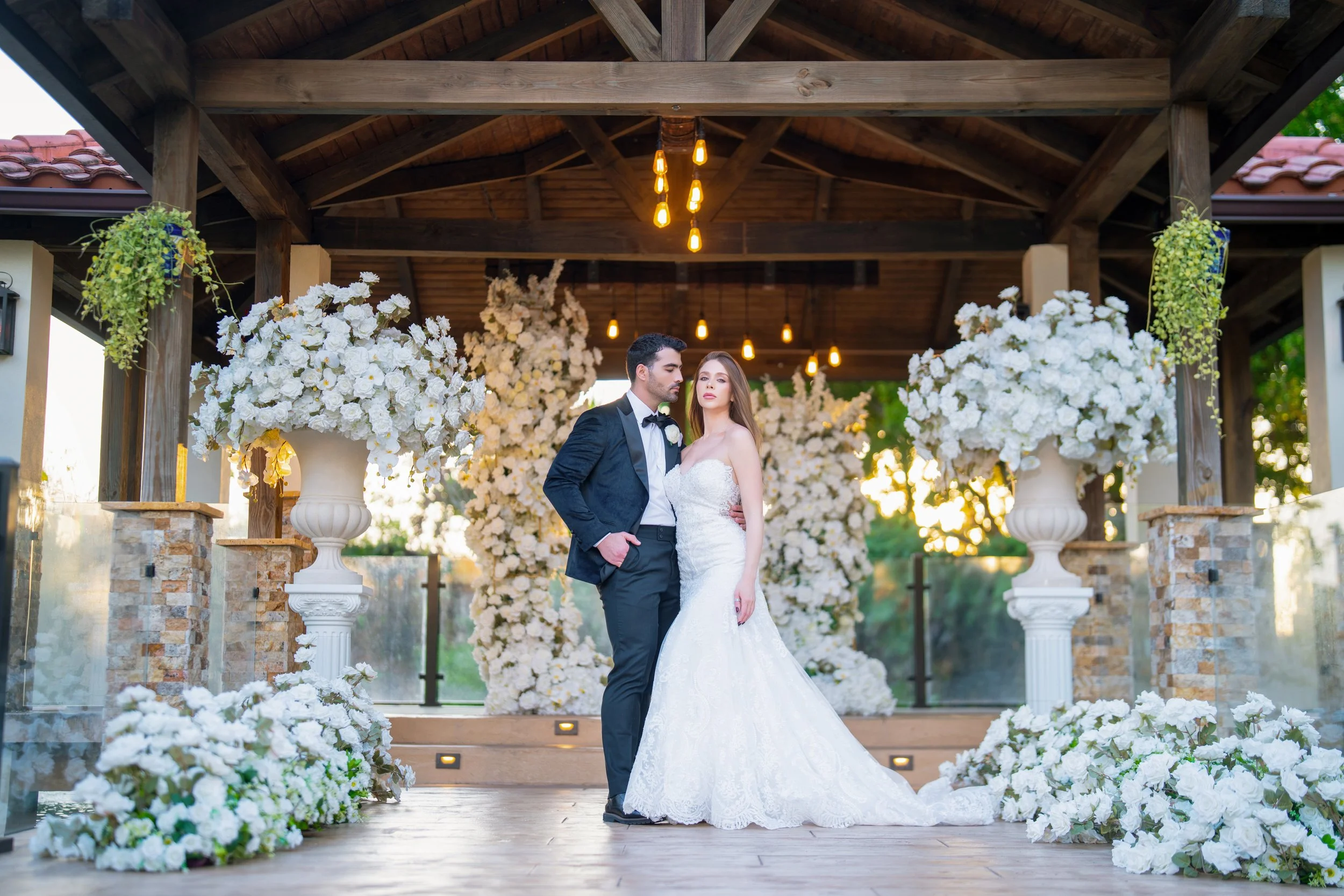 A bride and groom posing at their wedding ceremony under a wooden pavilion decorated with white flowers and large floral arrangements.