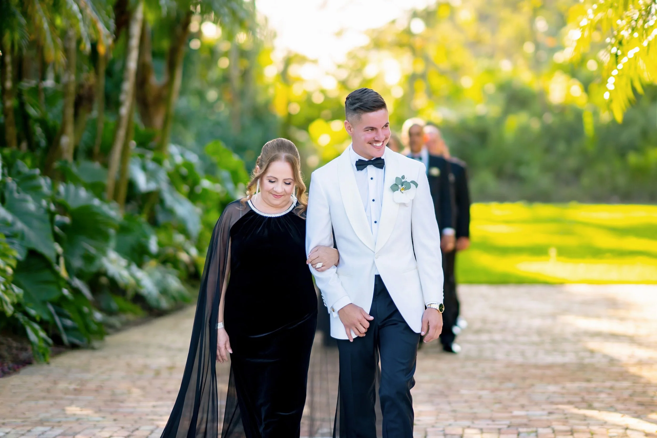 A couple in formal attire walking outdoors, the man in a white tuxedo jacket with a black bow tie and the woman in a long black velvet dress with sheer sleeves, smiling on a paved pathway surrounded by lush greenery and trees.