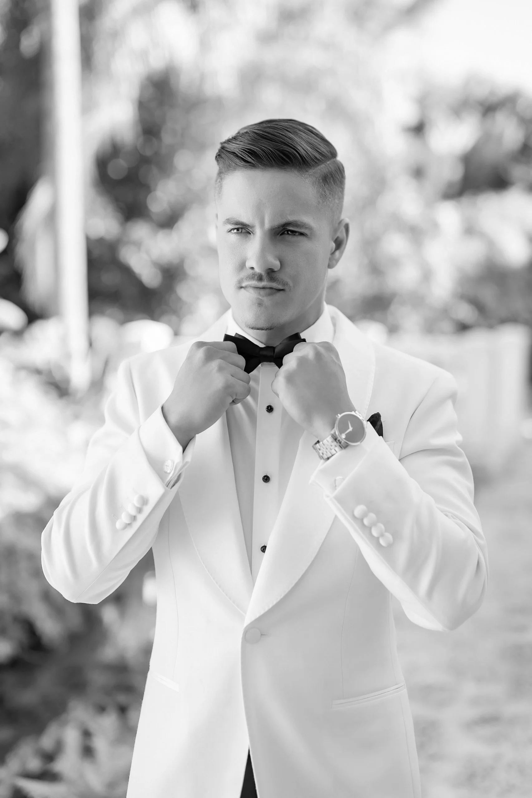 A young man in a tuxedo adjusting his bow tie outdoors, looking confident and focused.