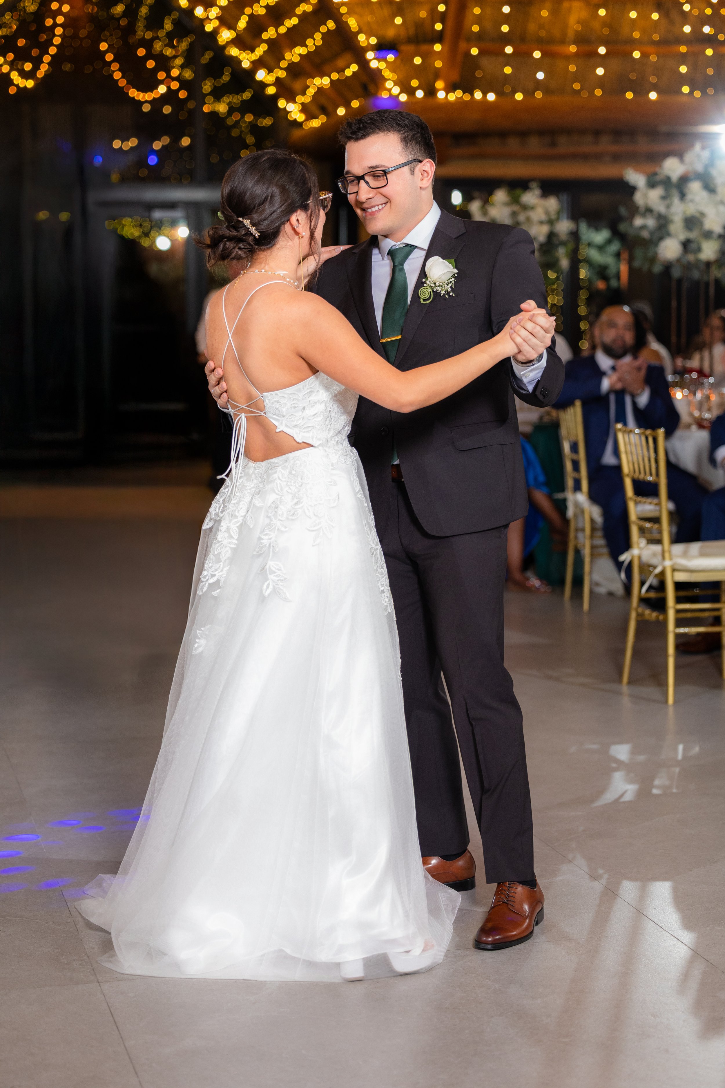 A bride and groom sharing their first dance at their wedding reception, with guests seated in the background and decorative string lights overhead.